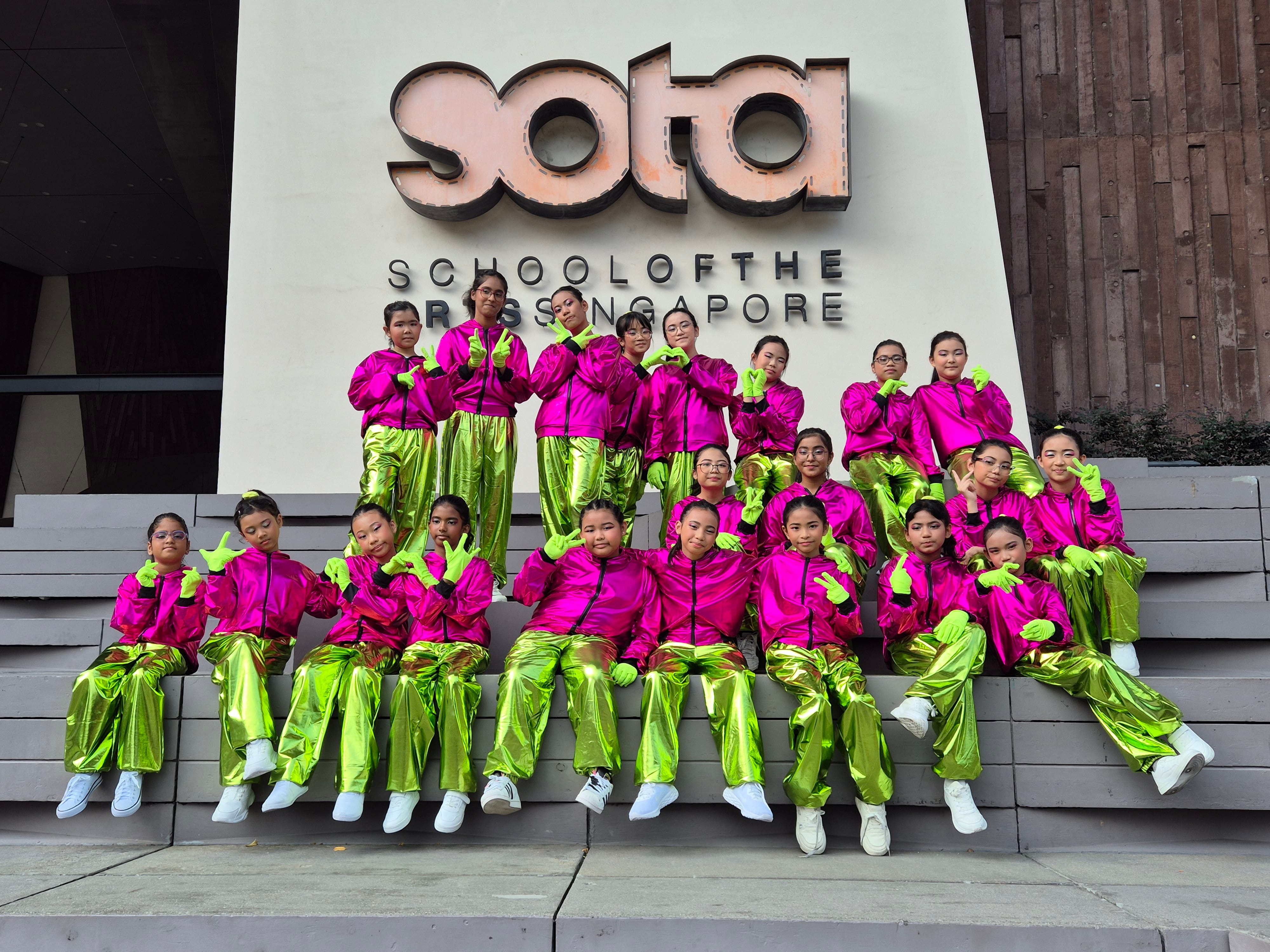 A group of young dancers in bright pink jackets and neon green pants pose in front of the SOTA Singapore sign.