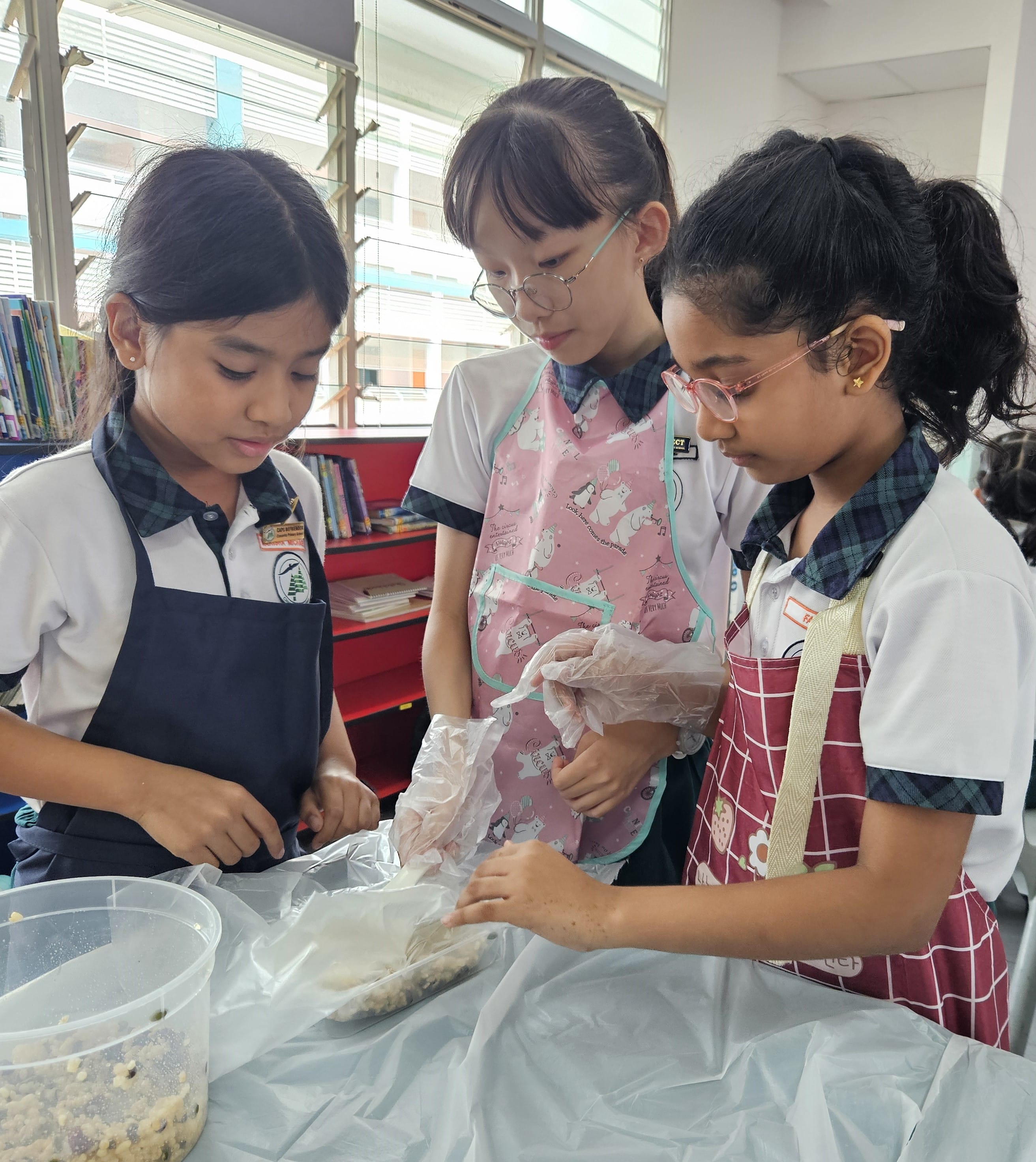 Three schoolgirls wearing aprons and gloves work together on a food preparation activity at a table.