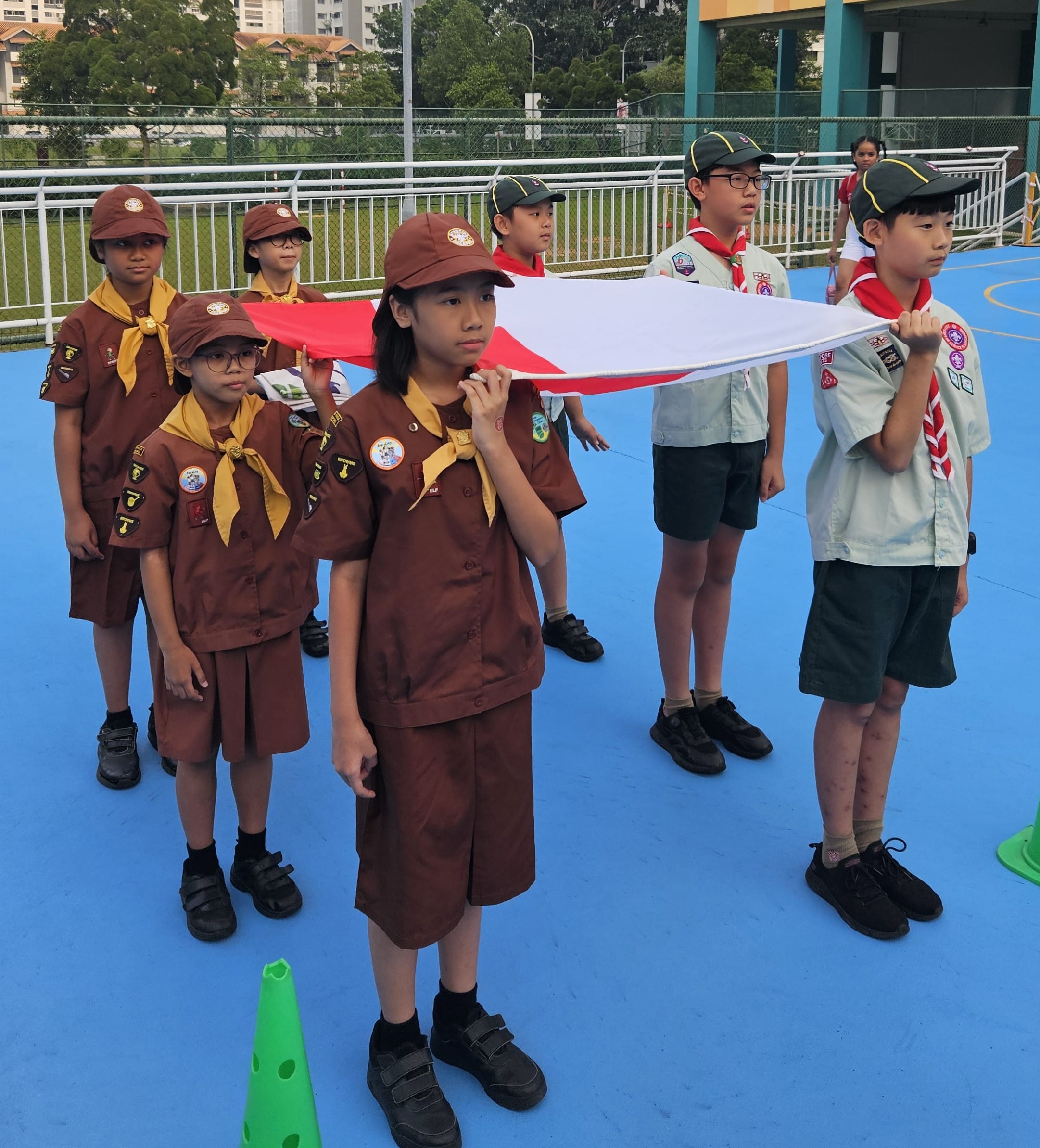 Children in scout uniforms hold a red and white flag on a blue outdoor court during a ceremony.