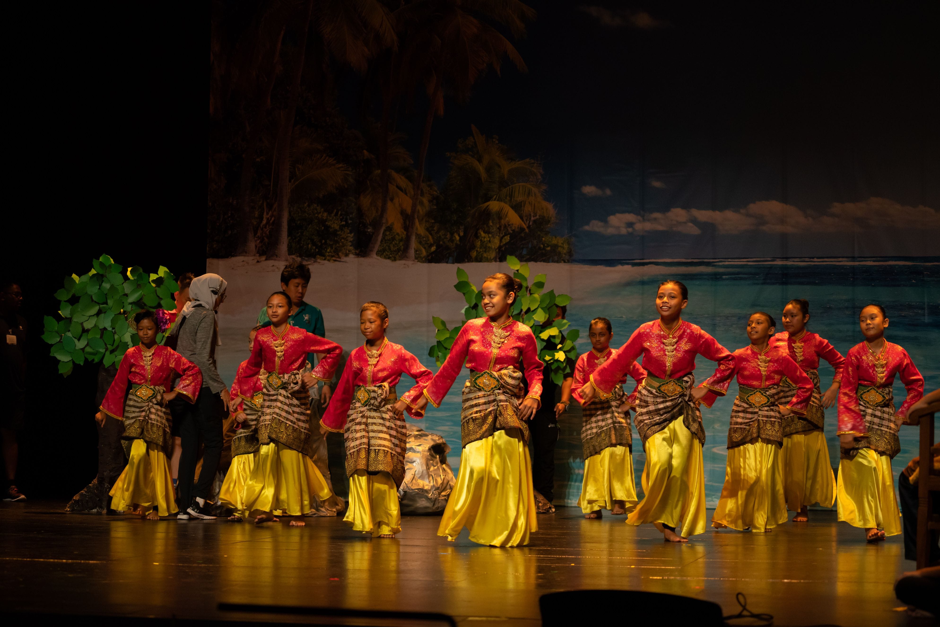 Girls in red and gold traditional costumes perform a cultural dance on a stage with a tropical beach backdrop.