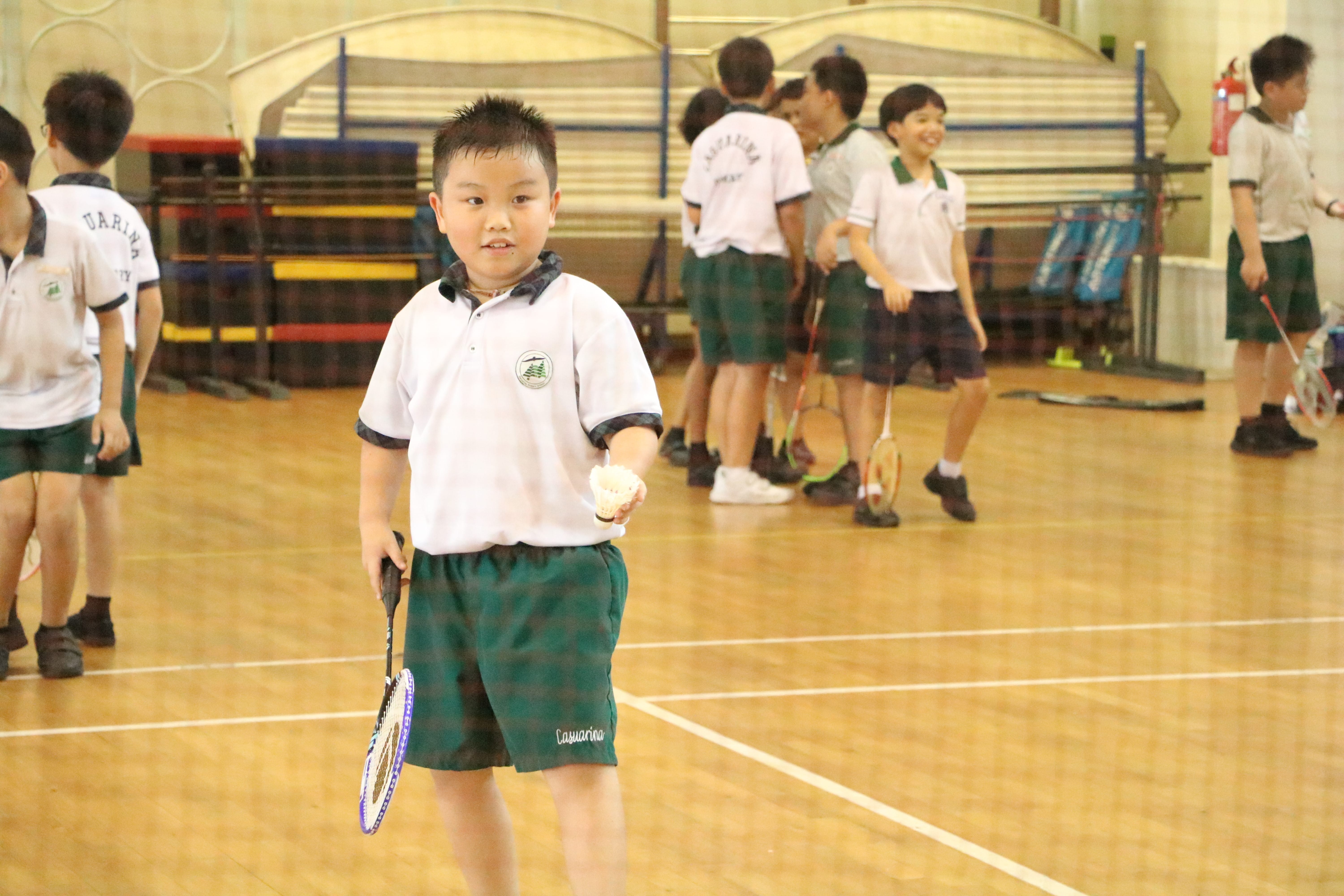 Schoolboy in gym holds a badminton racket and shuttlecock, with other students playing behind him.