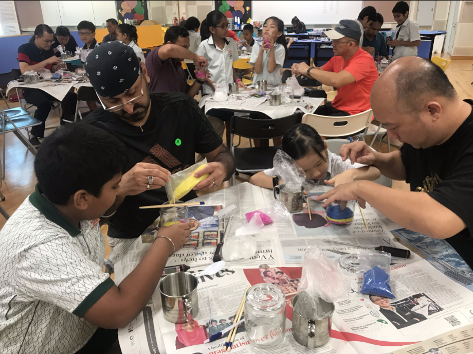 Adults and children sit at newspaper-covered tables doing a hands-on craft activity in a busy hall.