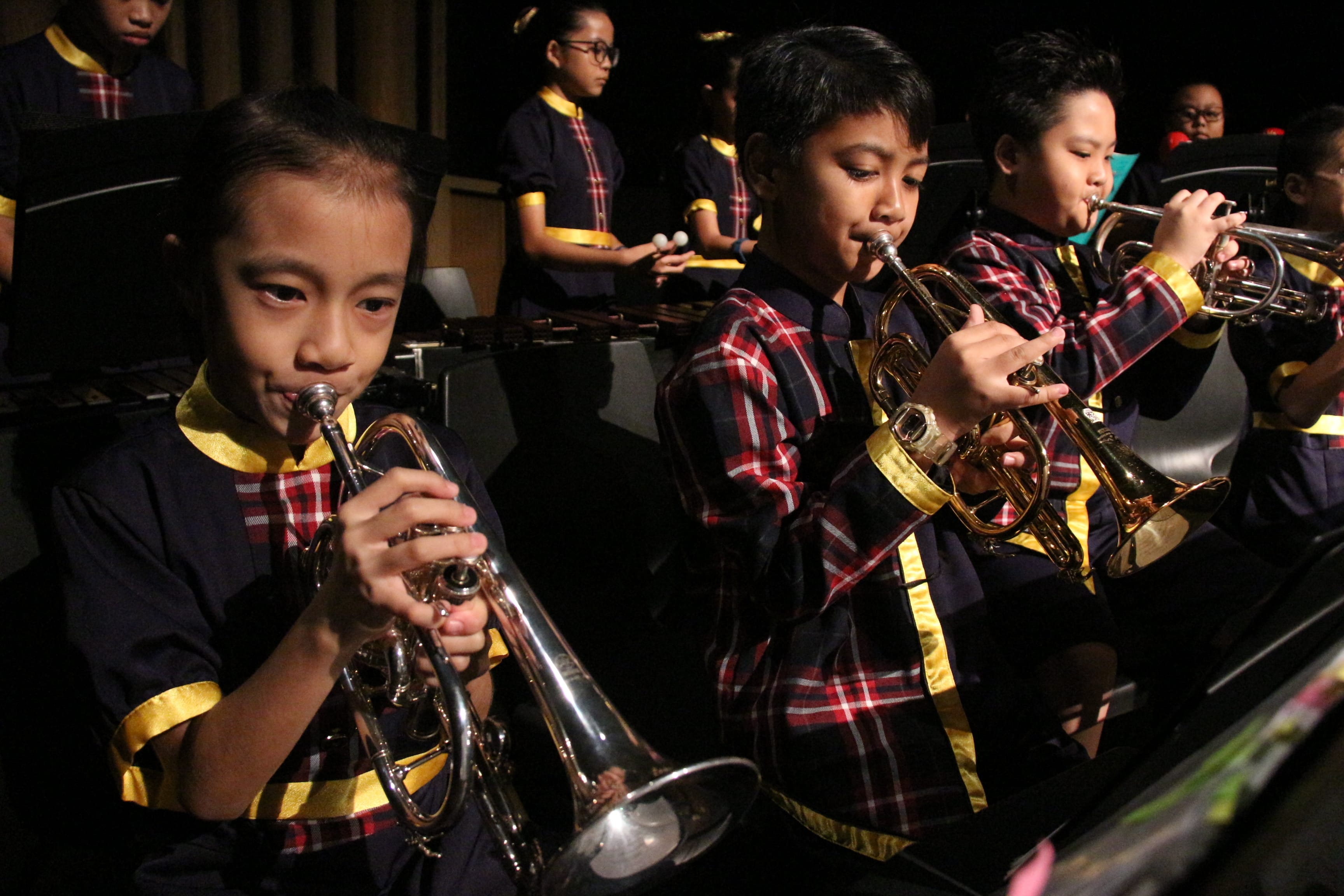 Children in matching plaid and navy uniforms play trumpets and percussion instruments in a youth band performance.