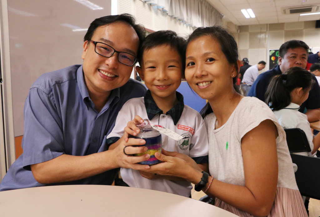 Two adults and a young boy smile together, holding a small jar filled with colorful layered sand.