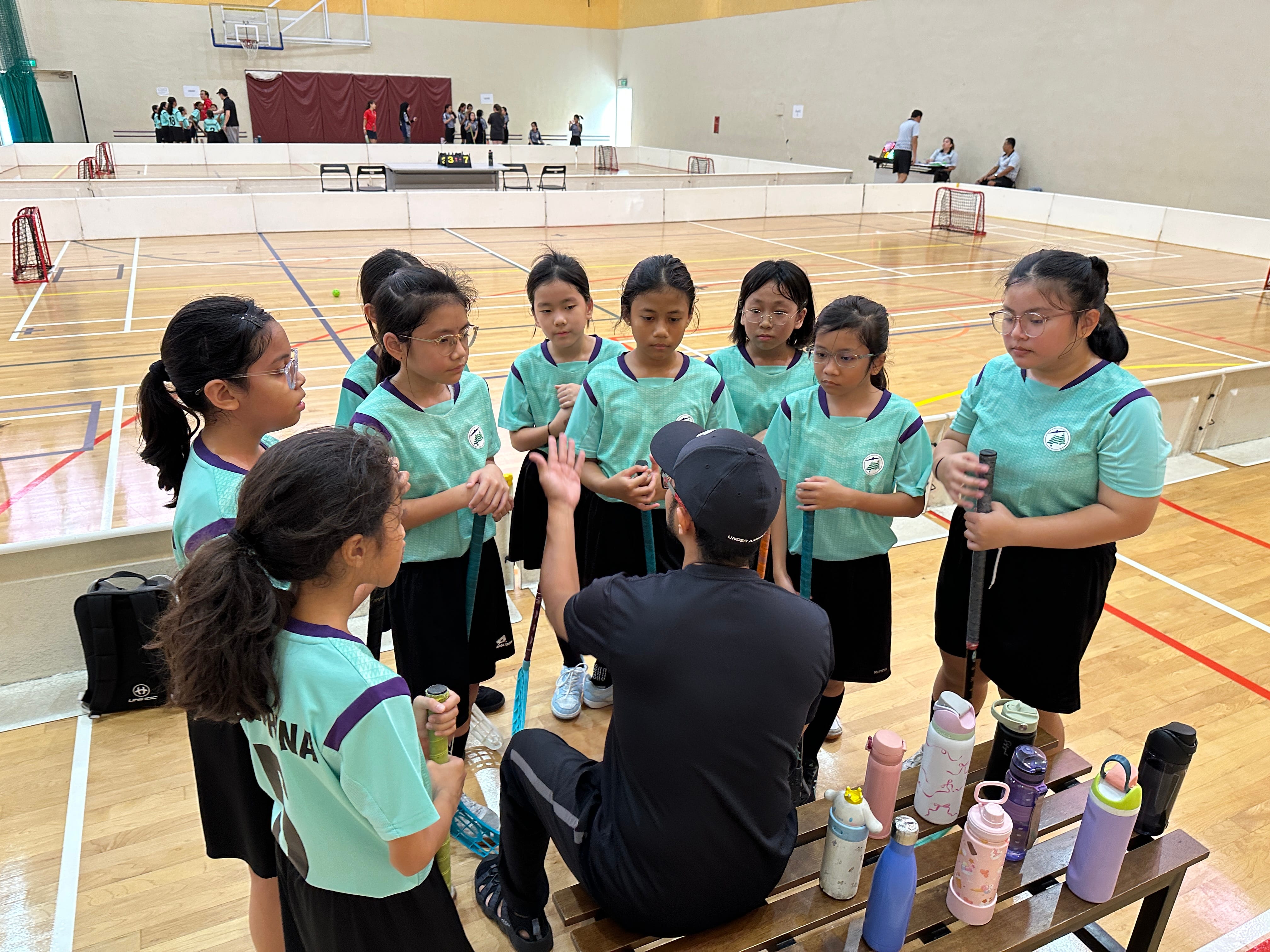 A coach in black huddles with a girls floorball team in teal jerseys inside a sports hall.