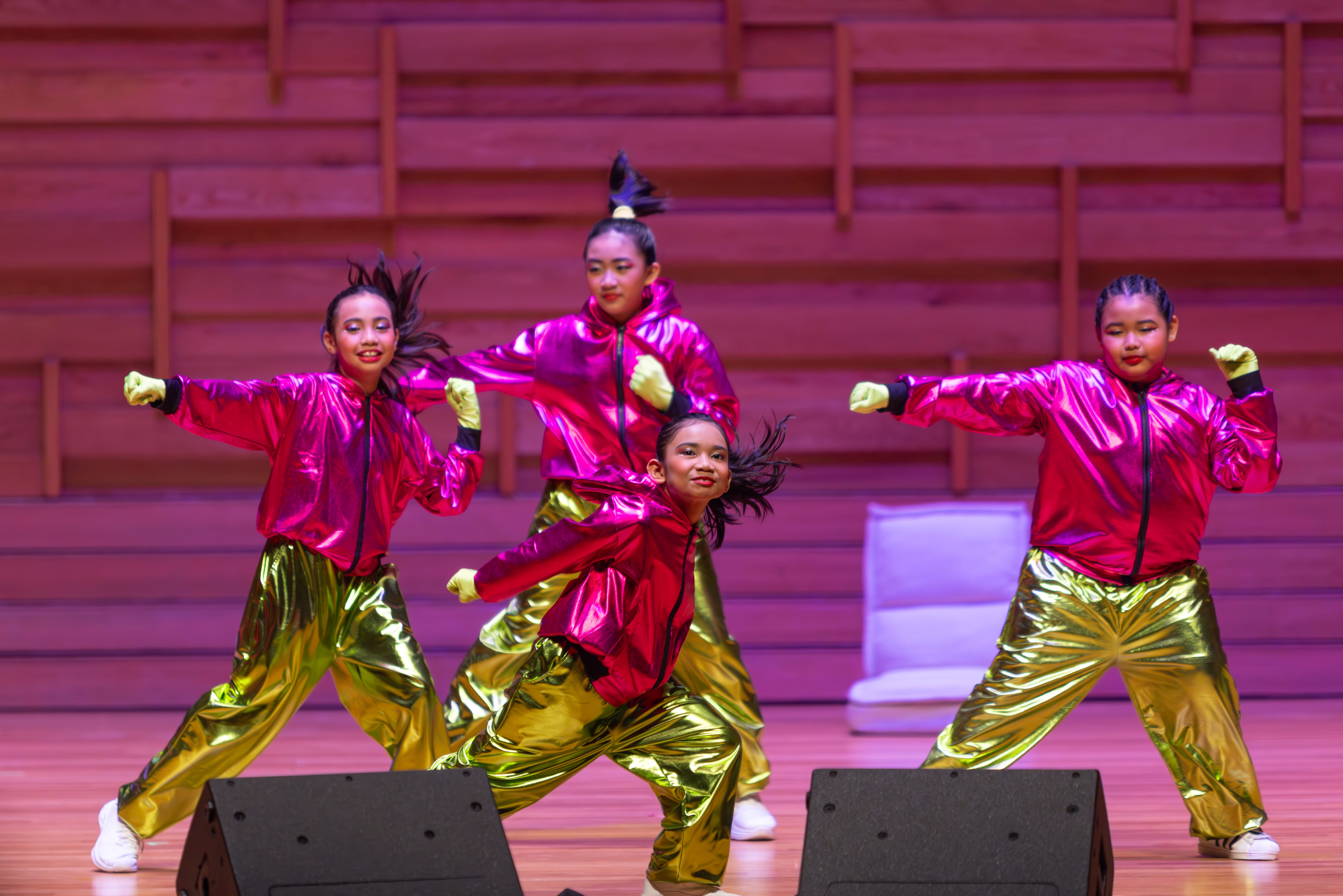 Four young girls perform hip-hop dance on stage wearing bright pink jackets and shiny gold pants.