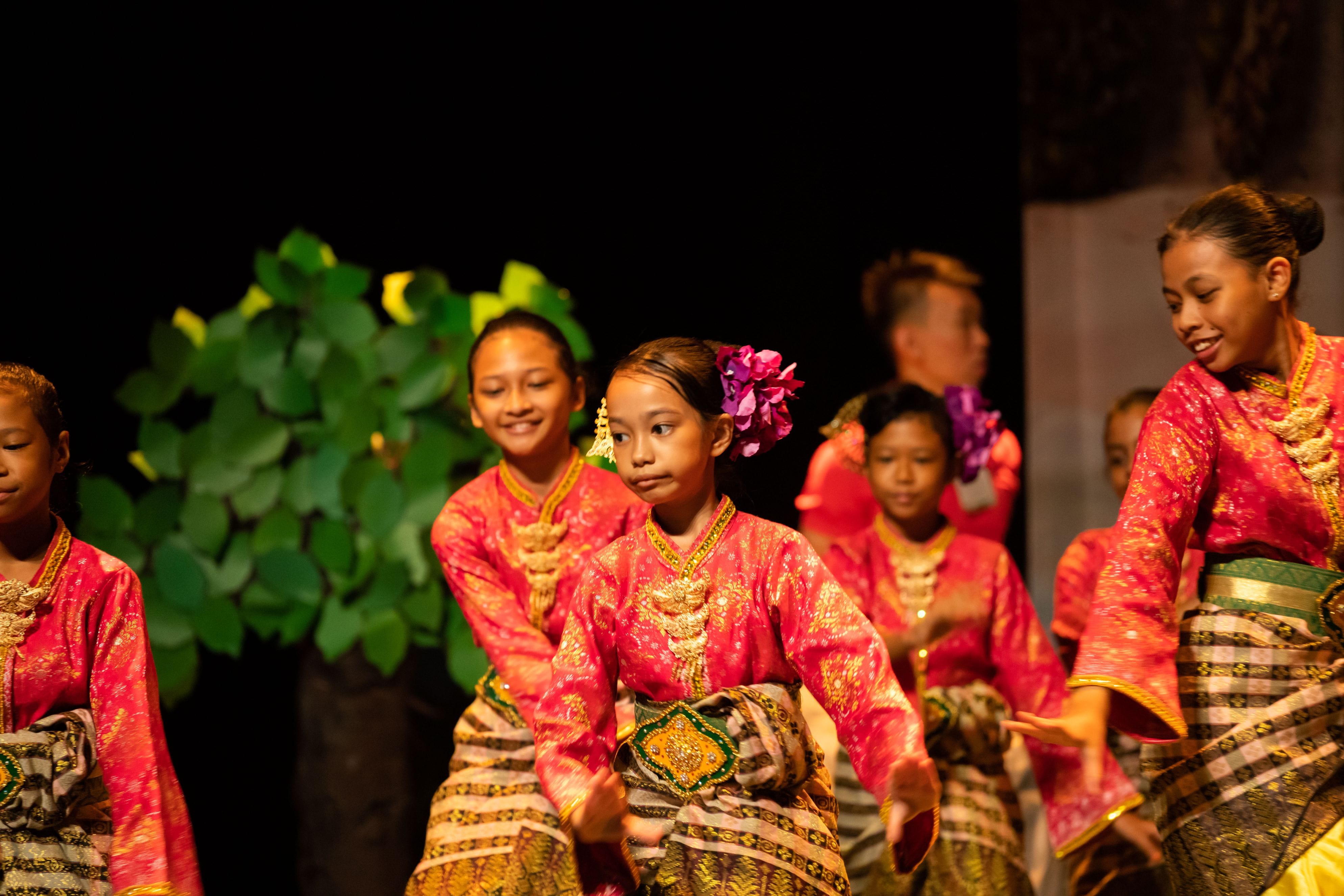 Young girls in red and gold traditional costumes with floral hair pieces perform a cultural dance on a dark stage.