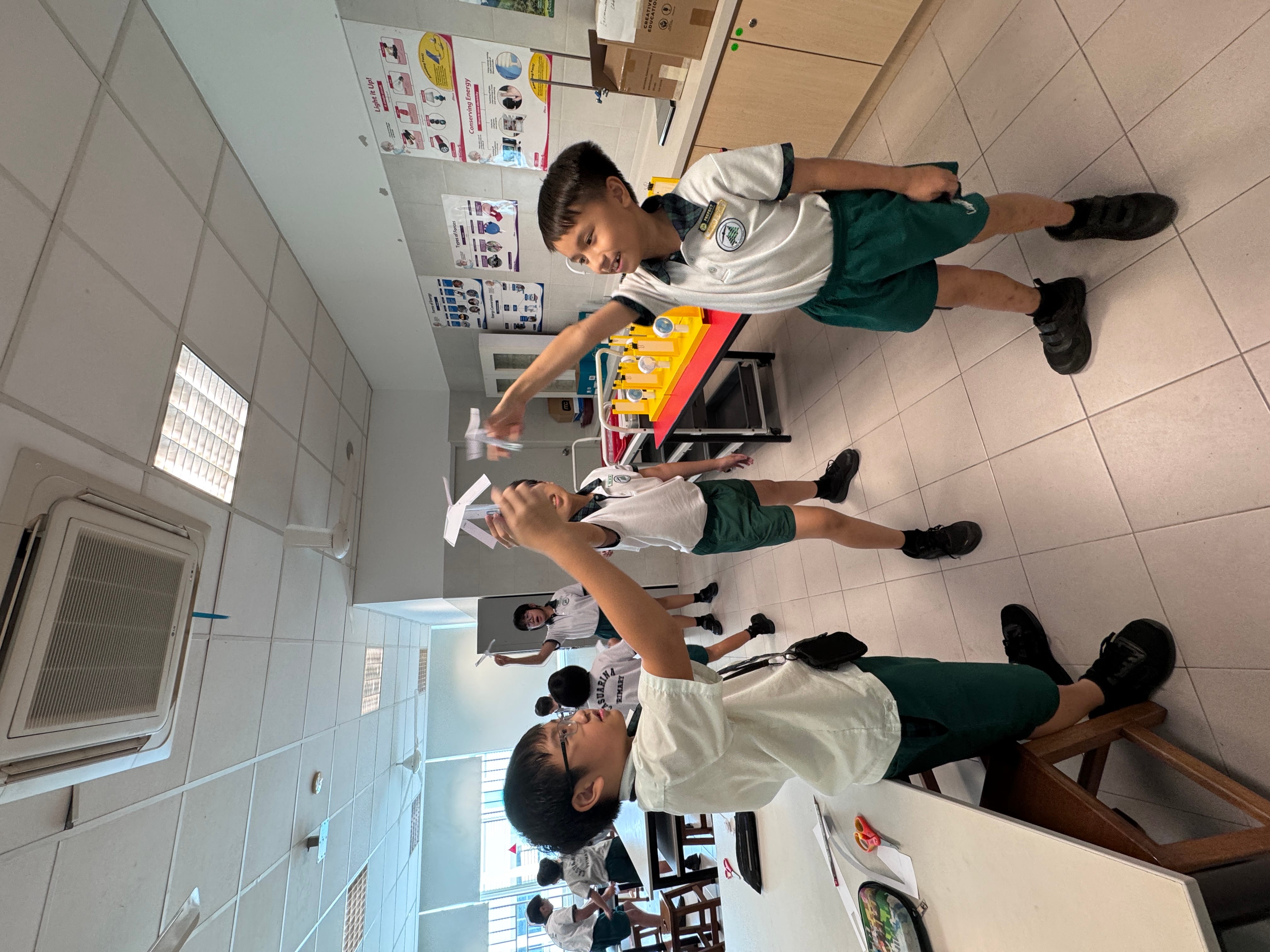 Three young students in white and green uniforms toss paper airplanes in a school science classroom.