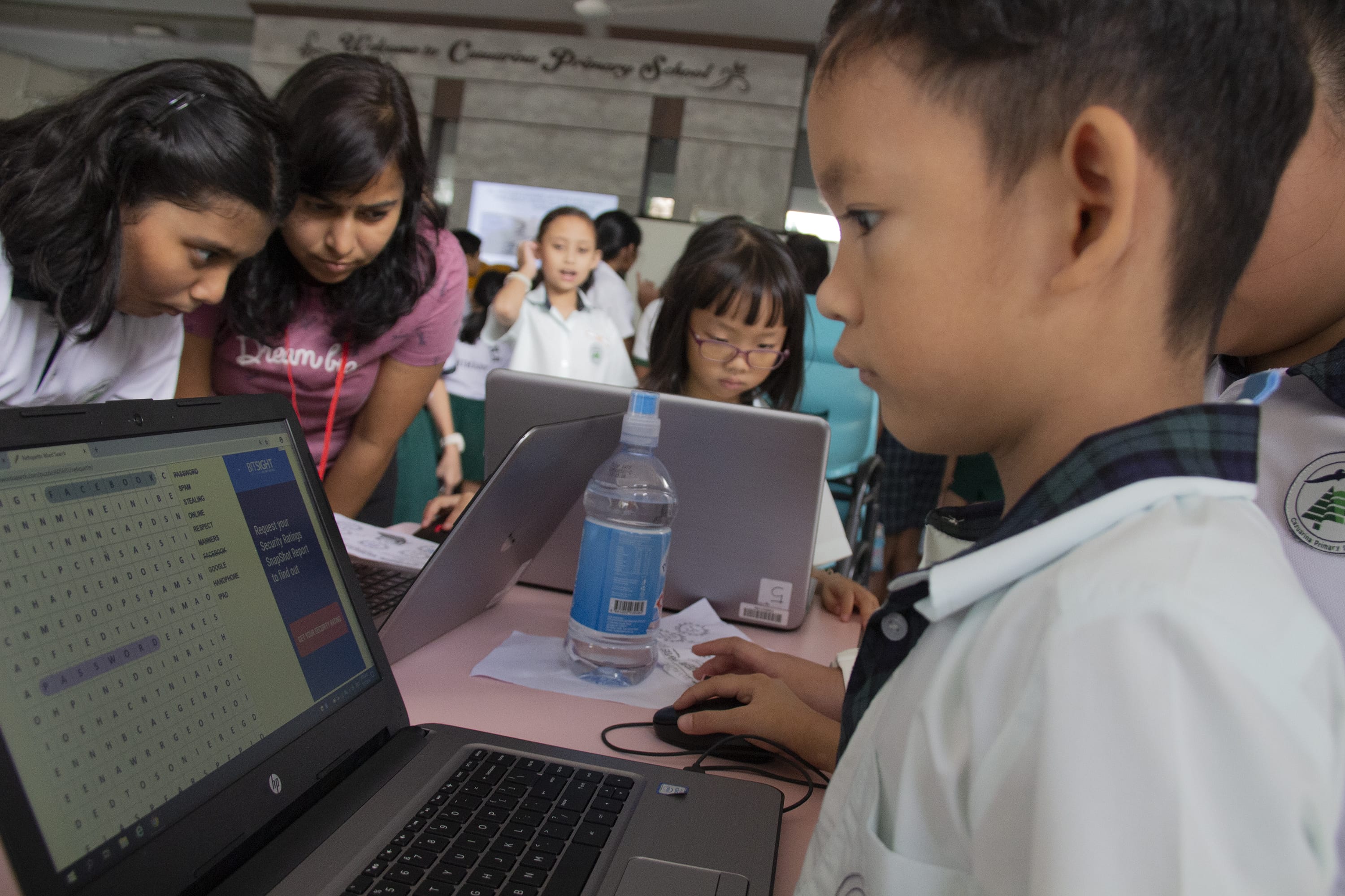 Schoolchildren and a teacher gathered around laptops on a table, one boy using a mouse in front.