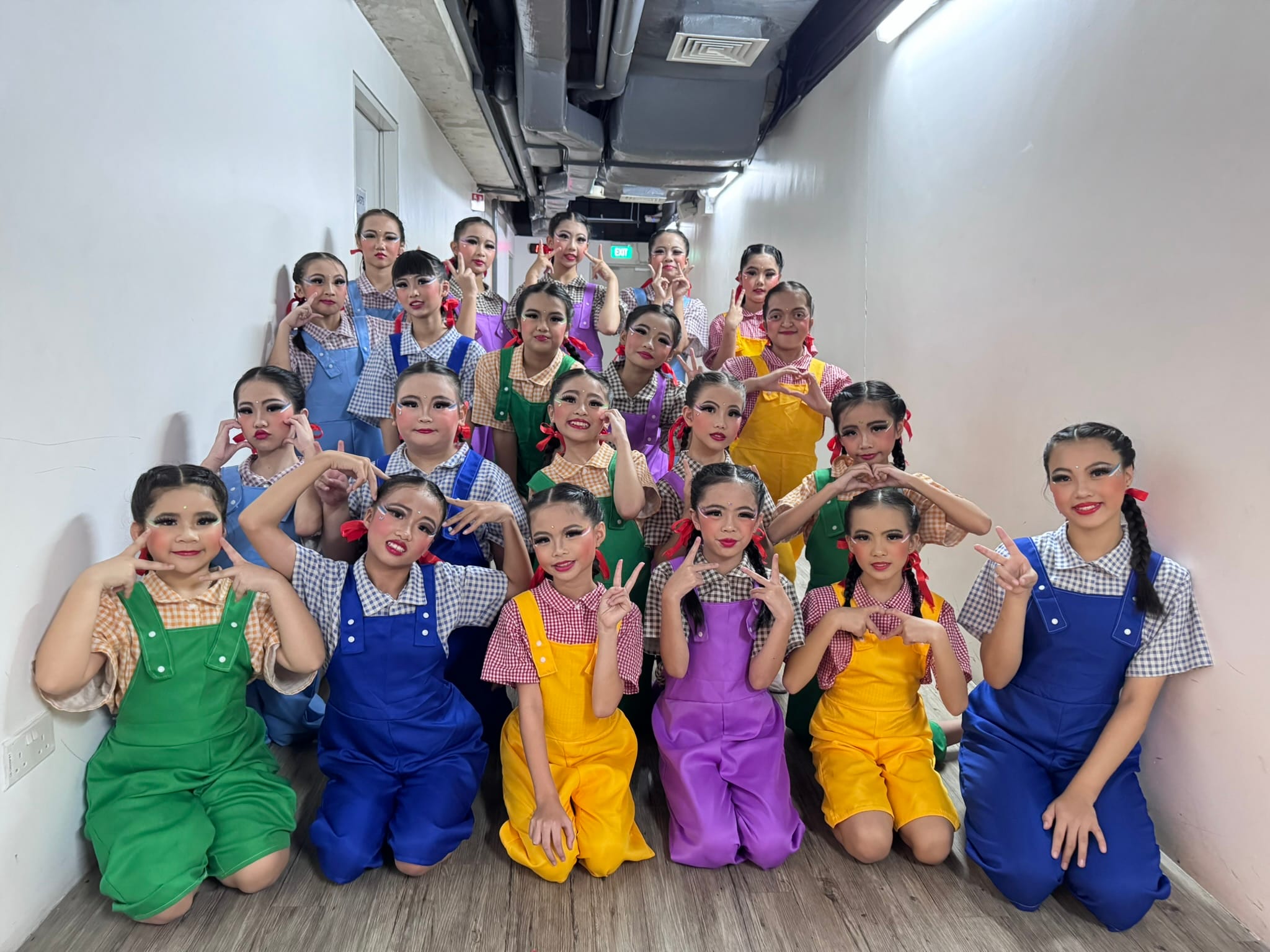 A large group of young girls in colorful overalls and checkered shirts with stage makeup pose in a hallway.