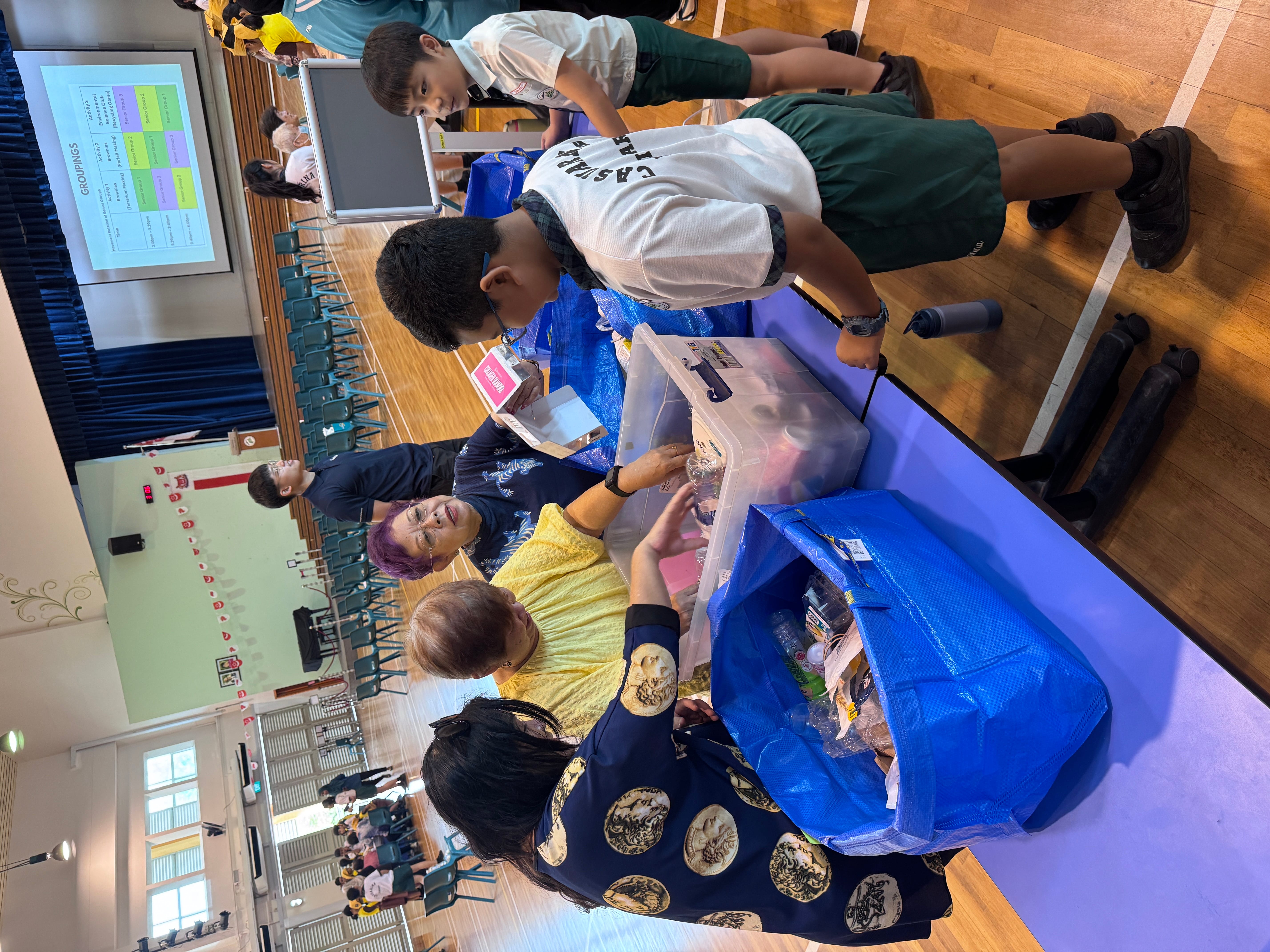 Two schoolboys lean over a table as three women sort items in a plastic bin and blue bag in a gym.