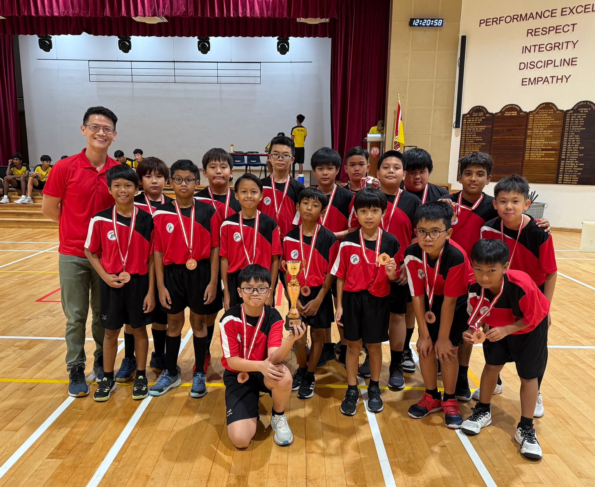A youth sports team in red and black uniforms poses with medals and a trophy in a school gymnasium.