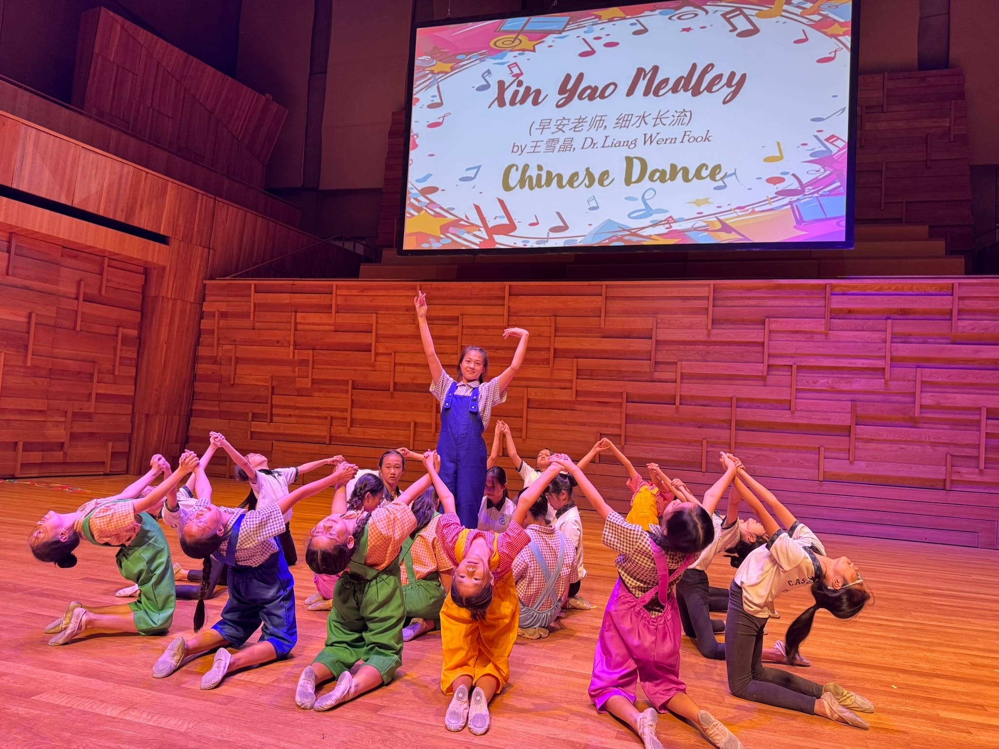 Children perform a Chinese dance on stage, with one girl standing arms raised while others kneel around her, screen reads "Xin Yao Medley."