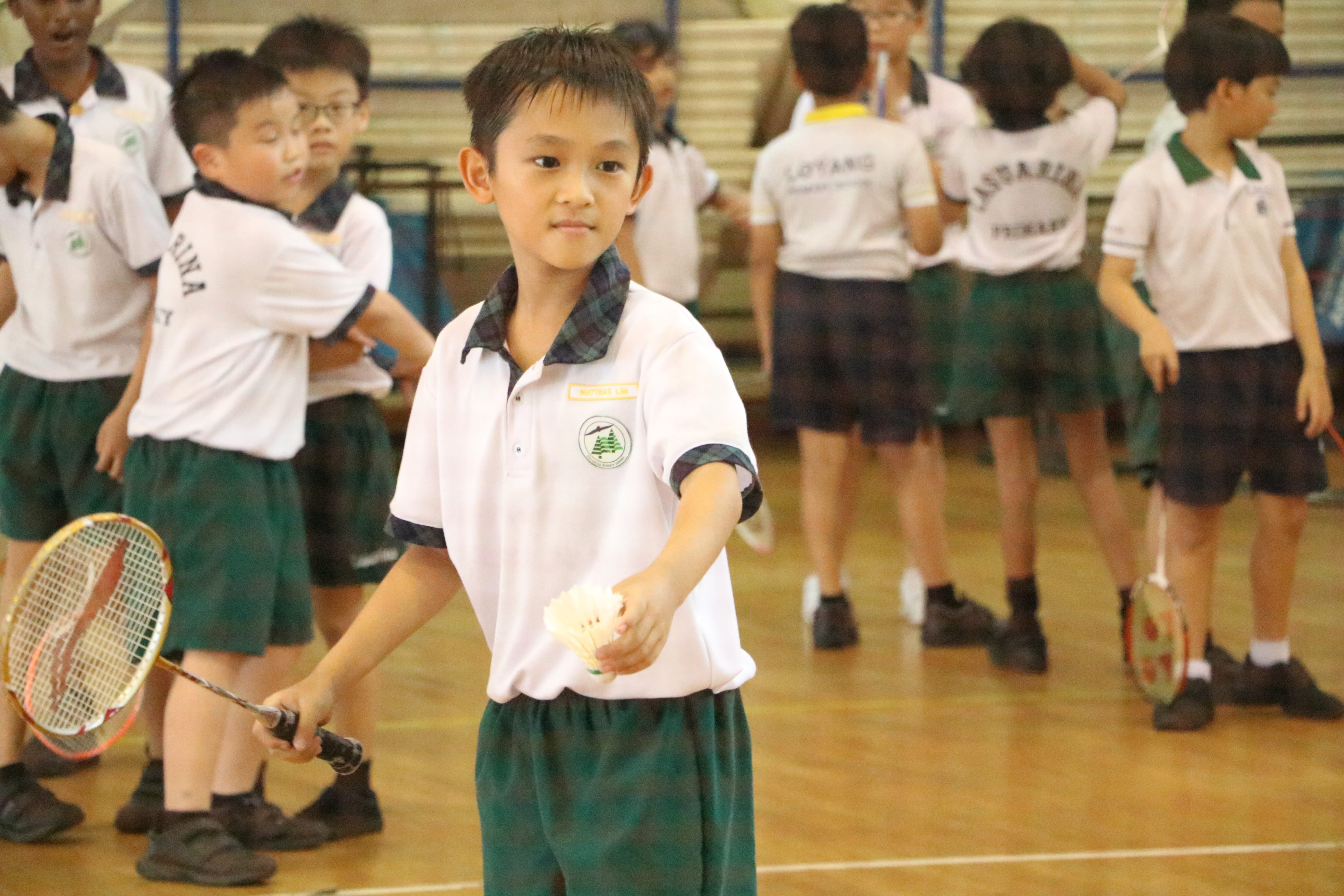 Schoolboy in uniform holds a badminton racket and shuttlecock in a gym with other students behind him