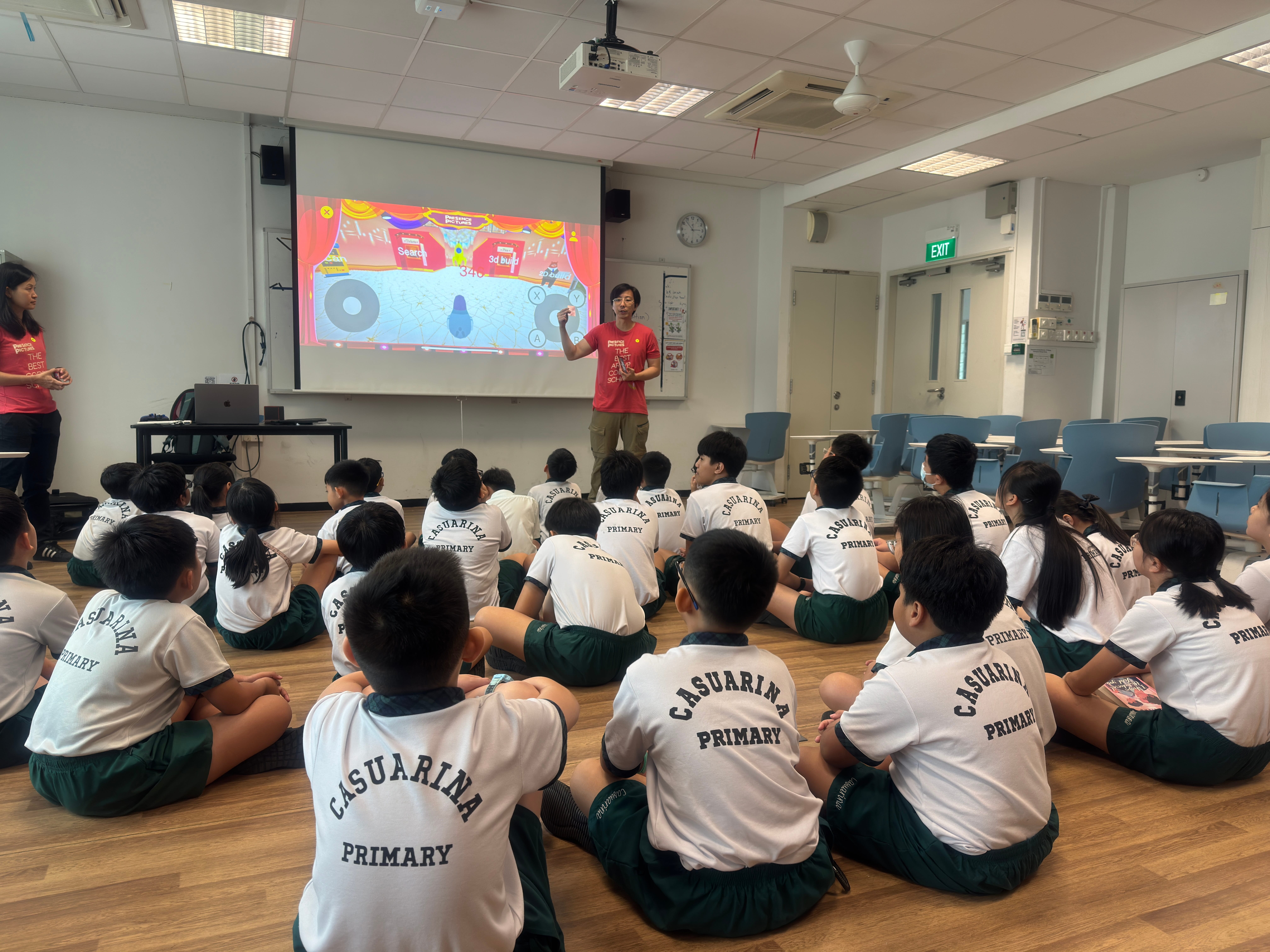 Children in school uniforms sit on floor facing teacher presenting colorful slide on projector screen.