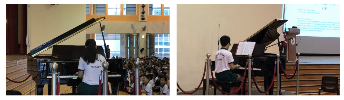 Two students in school uniforms play grand pianos on a stage before a seated audience in a school hall.