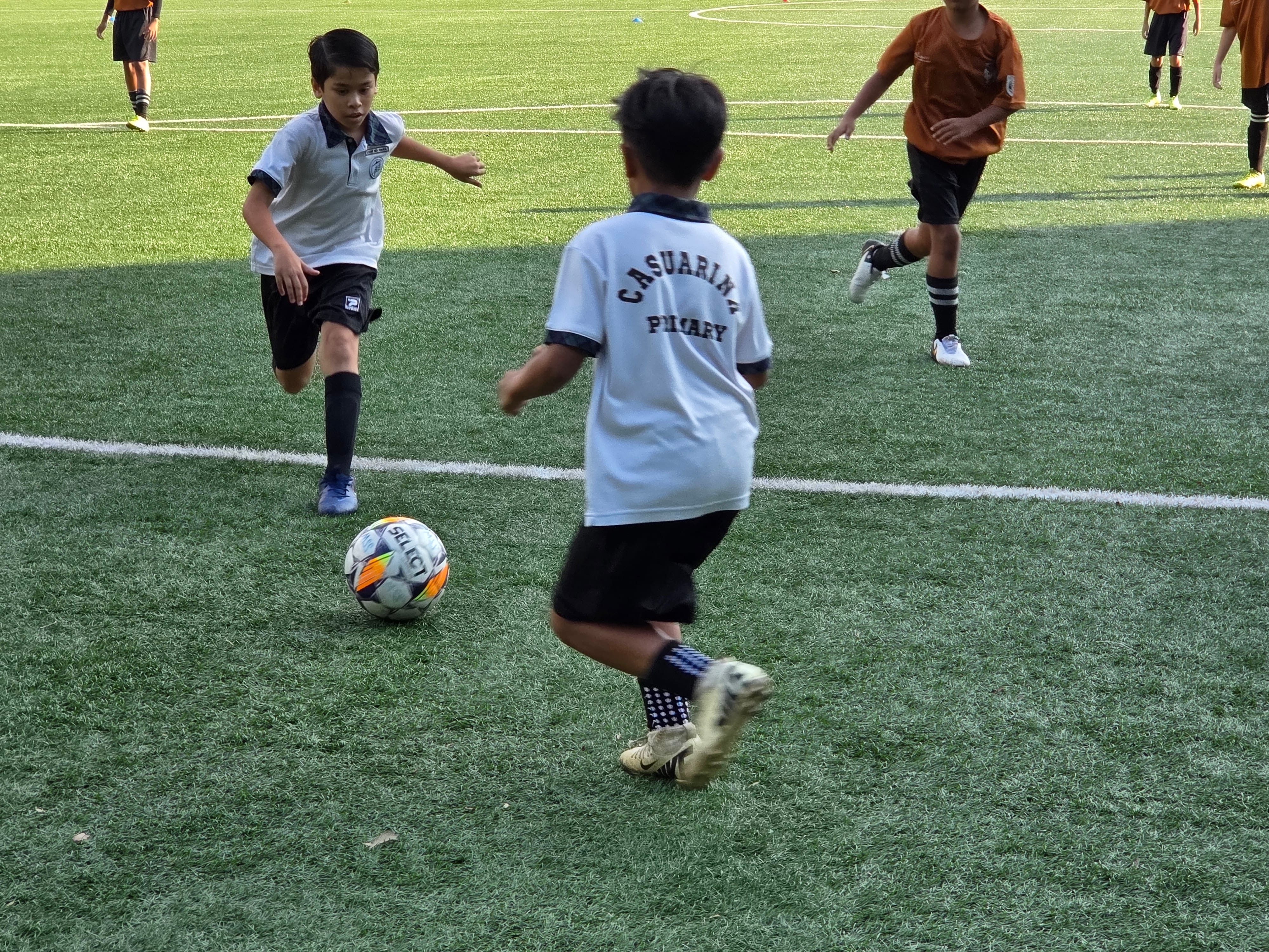 Two boys in white jerseys chase a soccer ball on a turf field while opponents in orange run nearby.