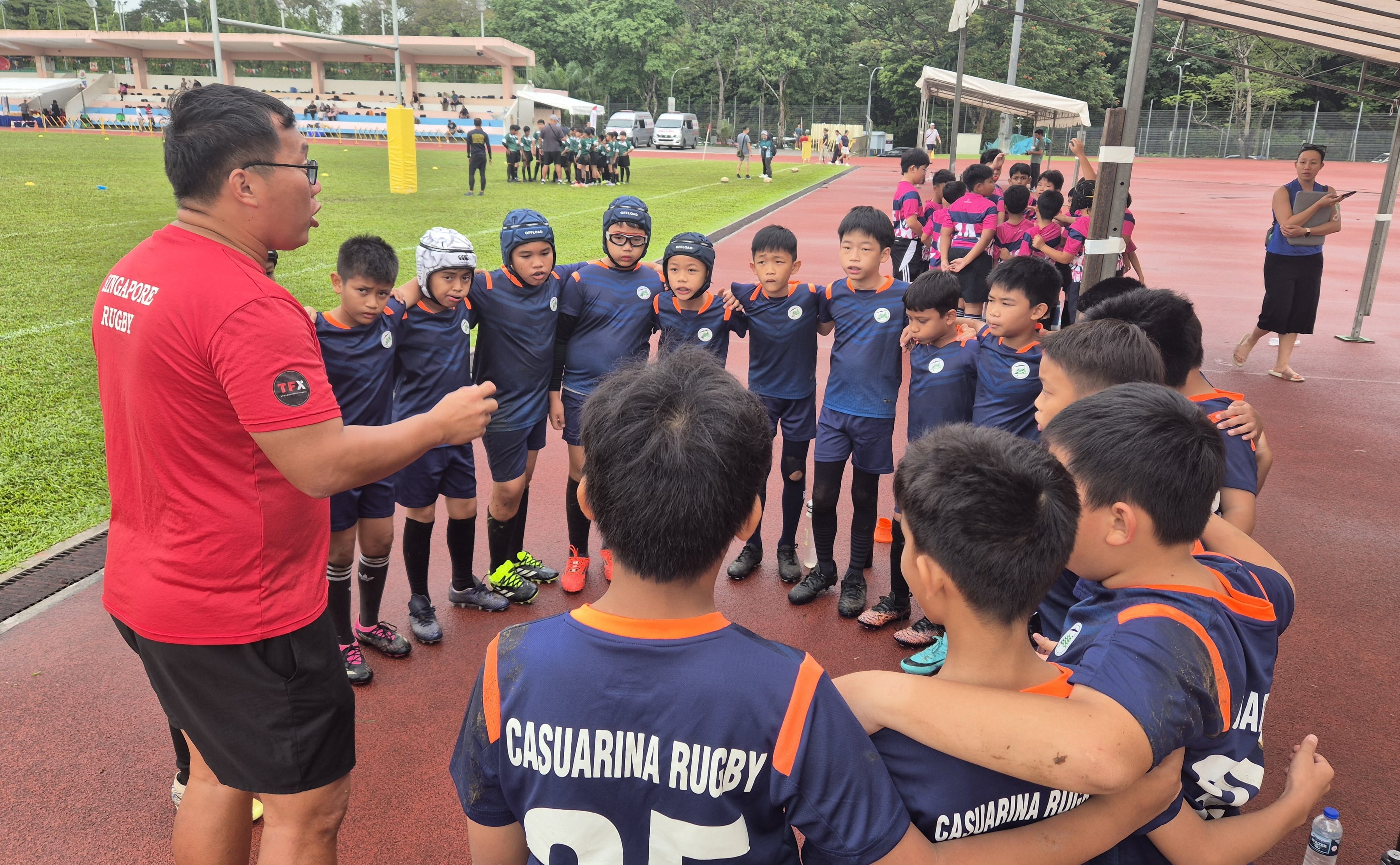 A Singapore Rugby coach in red addresses young Casuarina Rugby players in navy uniforms on a sports track.