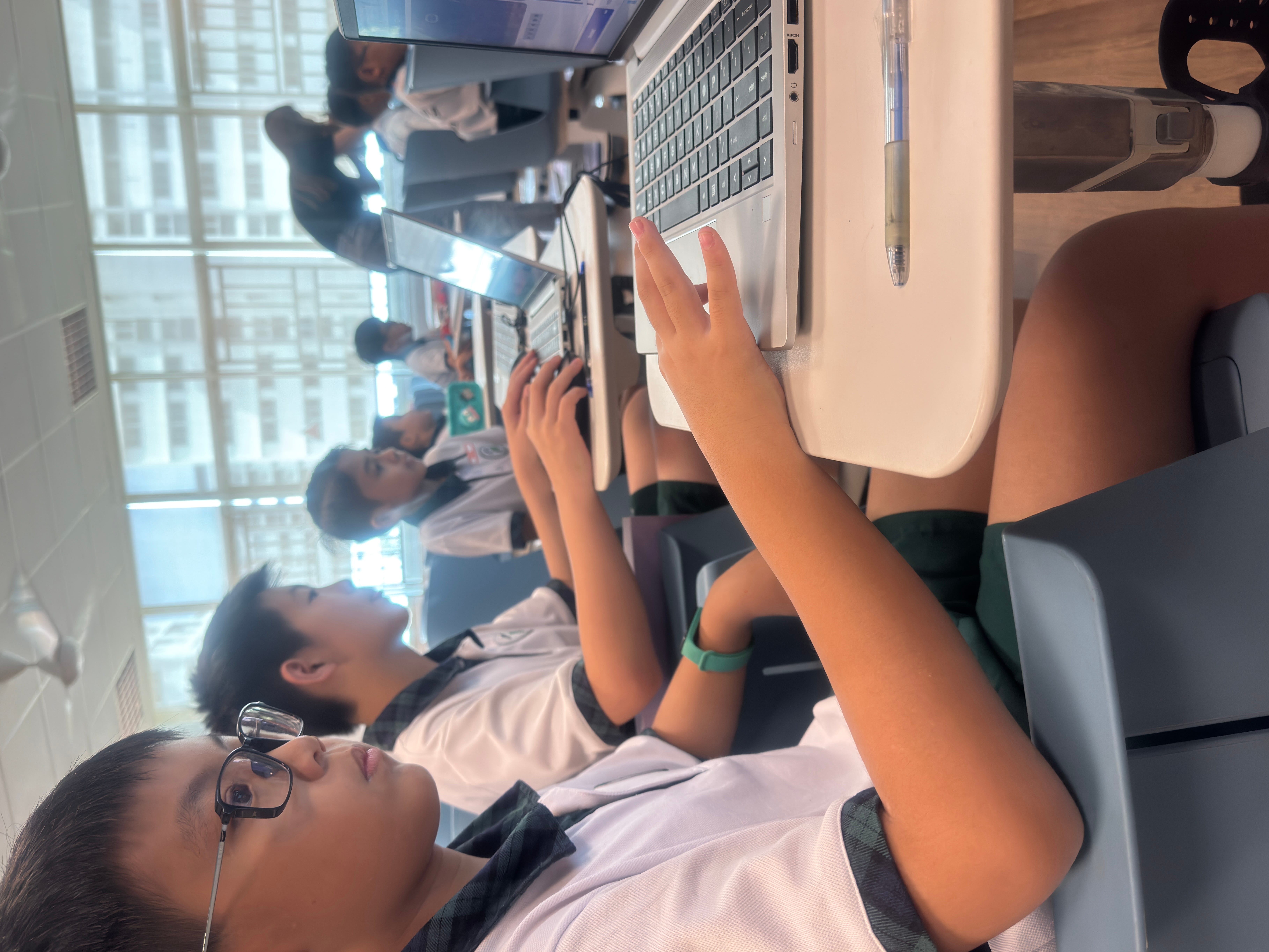 Students in uniform sit at desks using laptops; one wears glasses, with a pen on the desk nearby.