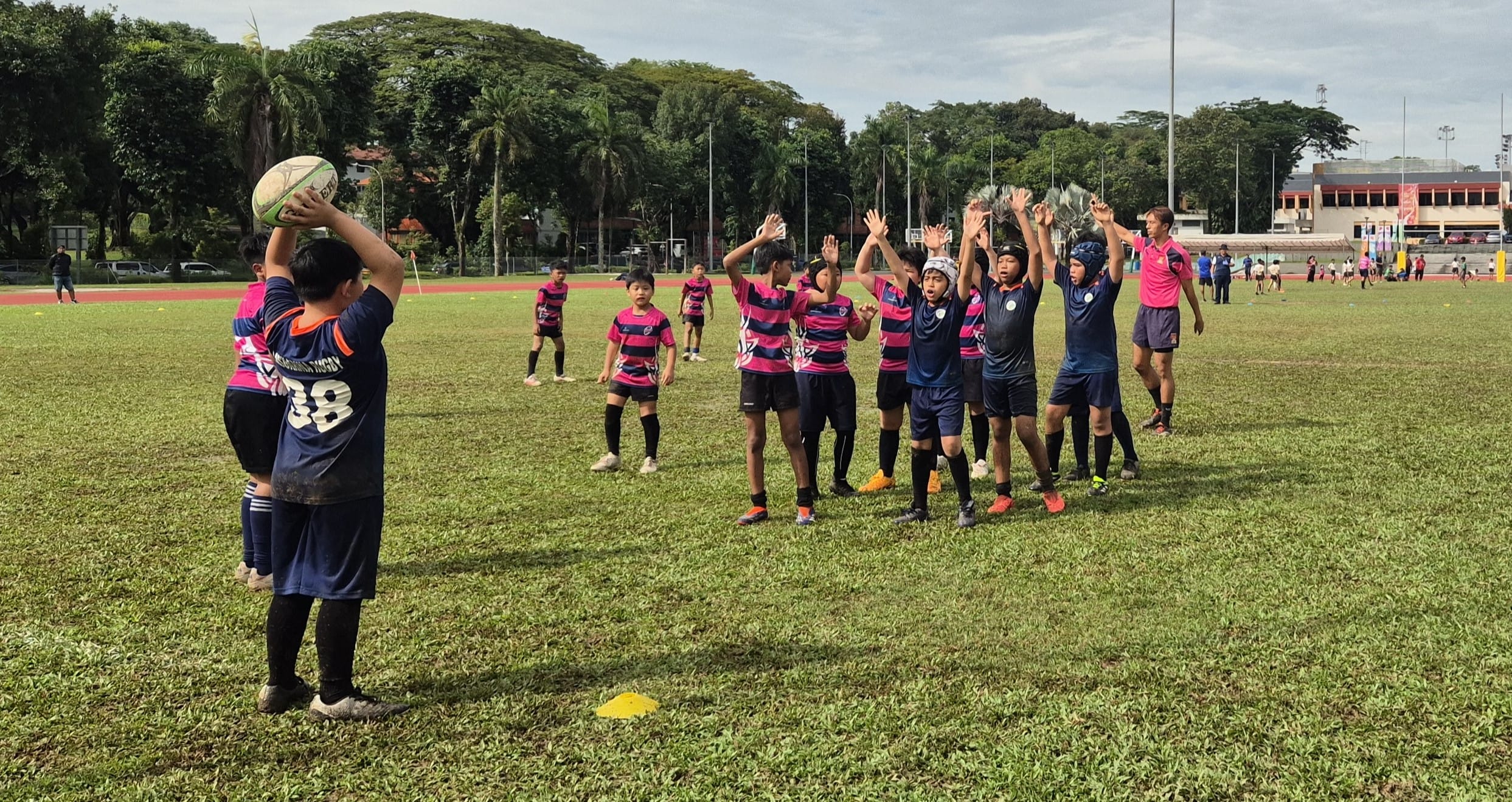 Children in pink and navy jerseys play rugby on a grass field, one throwing the ball as others reach up.