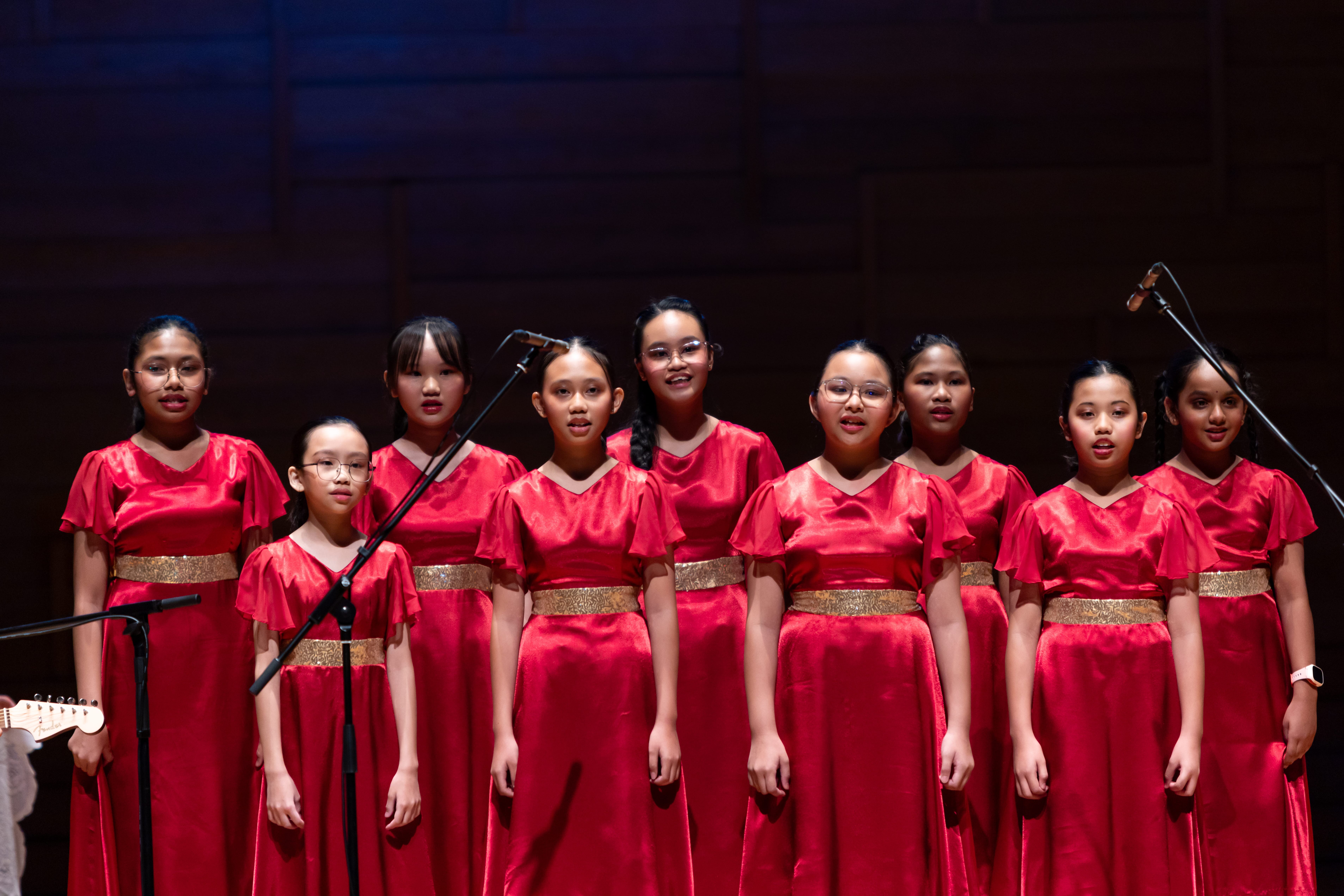 Nine girls in matching red satin dresses with gold belts singing on stage near microphone stands.