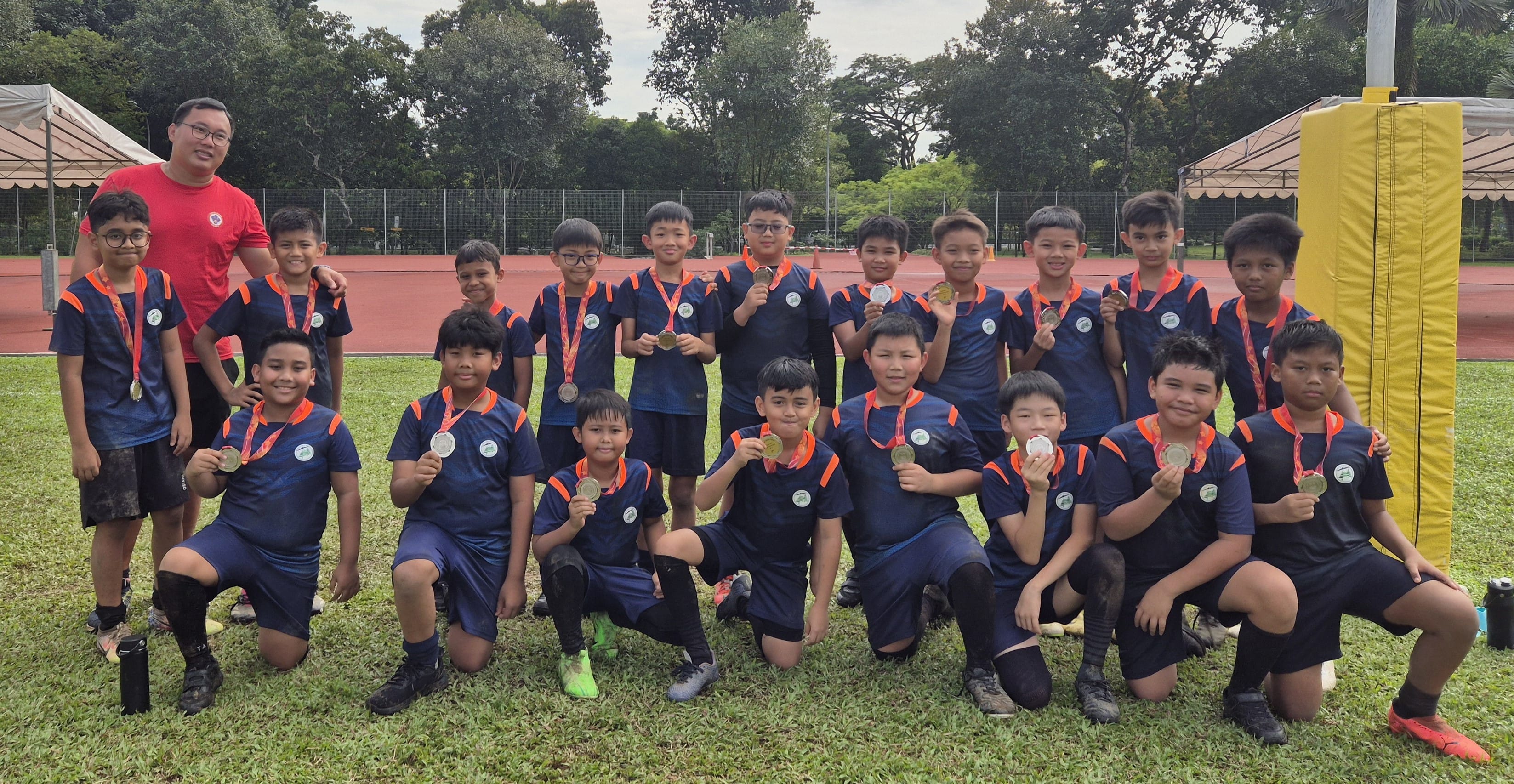 A youth football team in navy and orange jerseys poses with medals on a grass field beside a running track.