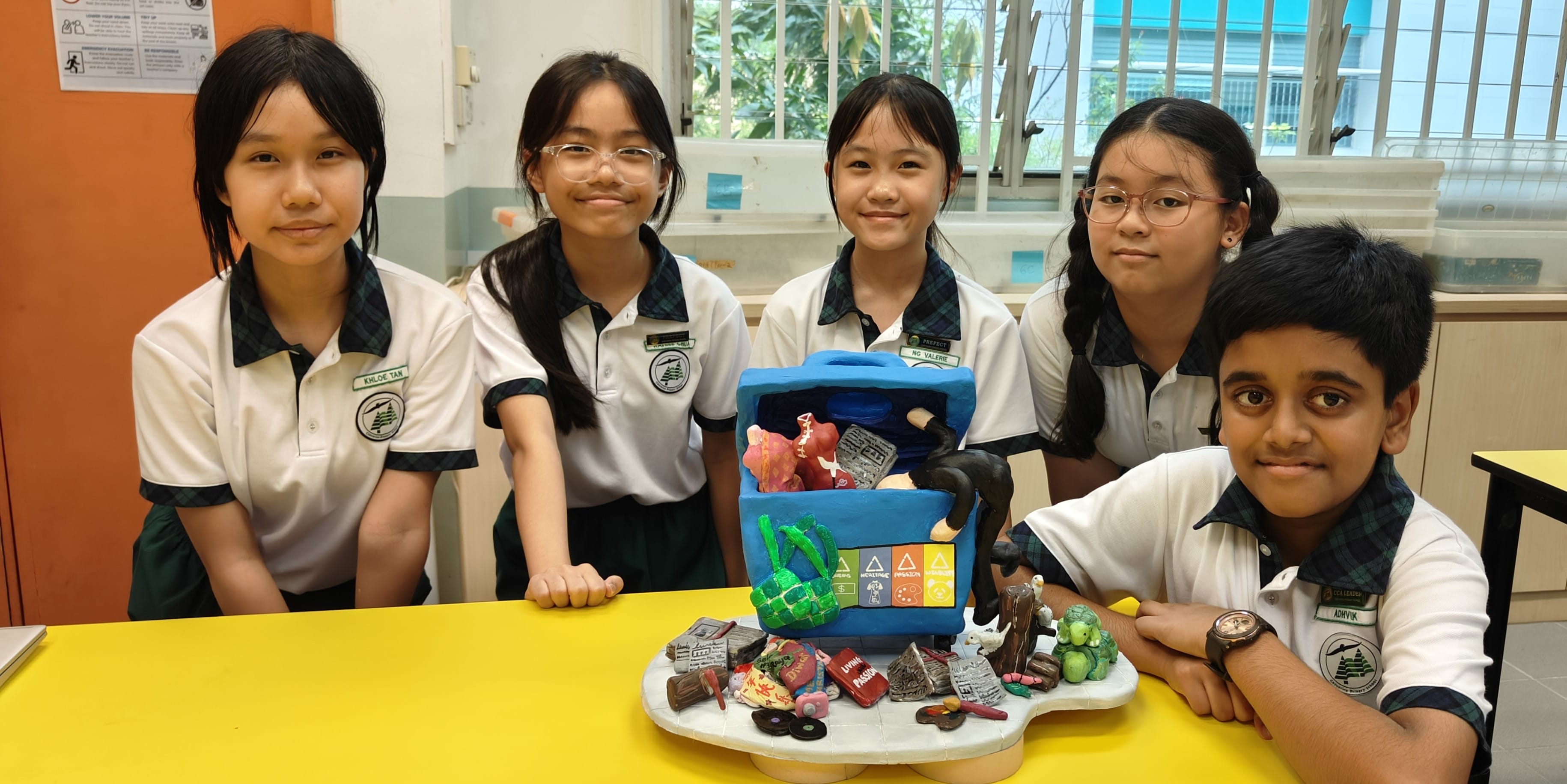 Five school students in uniform pose behind a yellow table displaying a colorful clay model project.