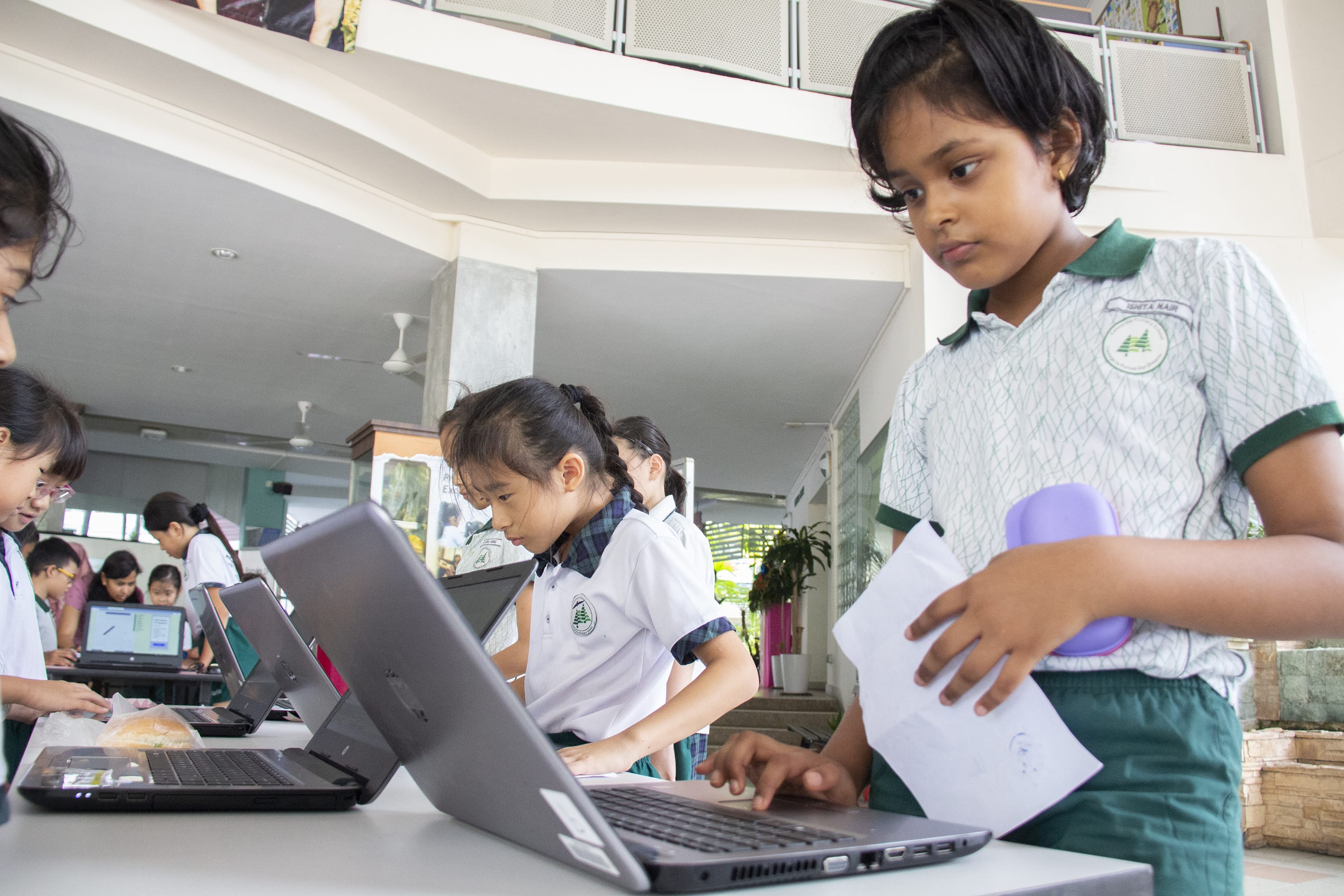 Schoolchildren in uniforms work at laptops on tables indoors; one girl holds papers and a purple bottle.