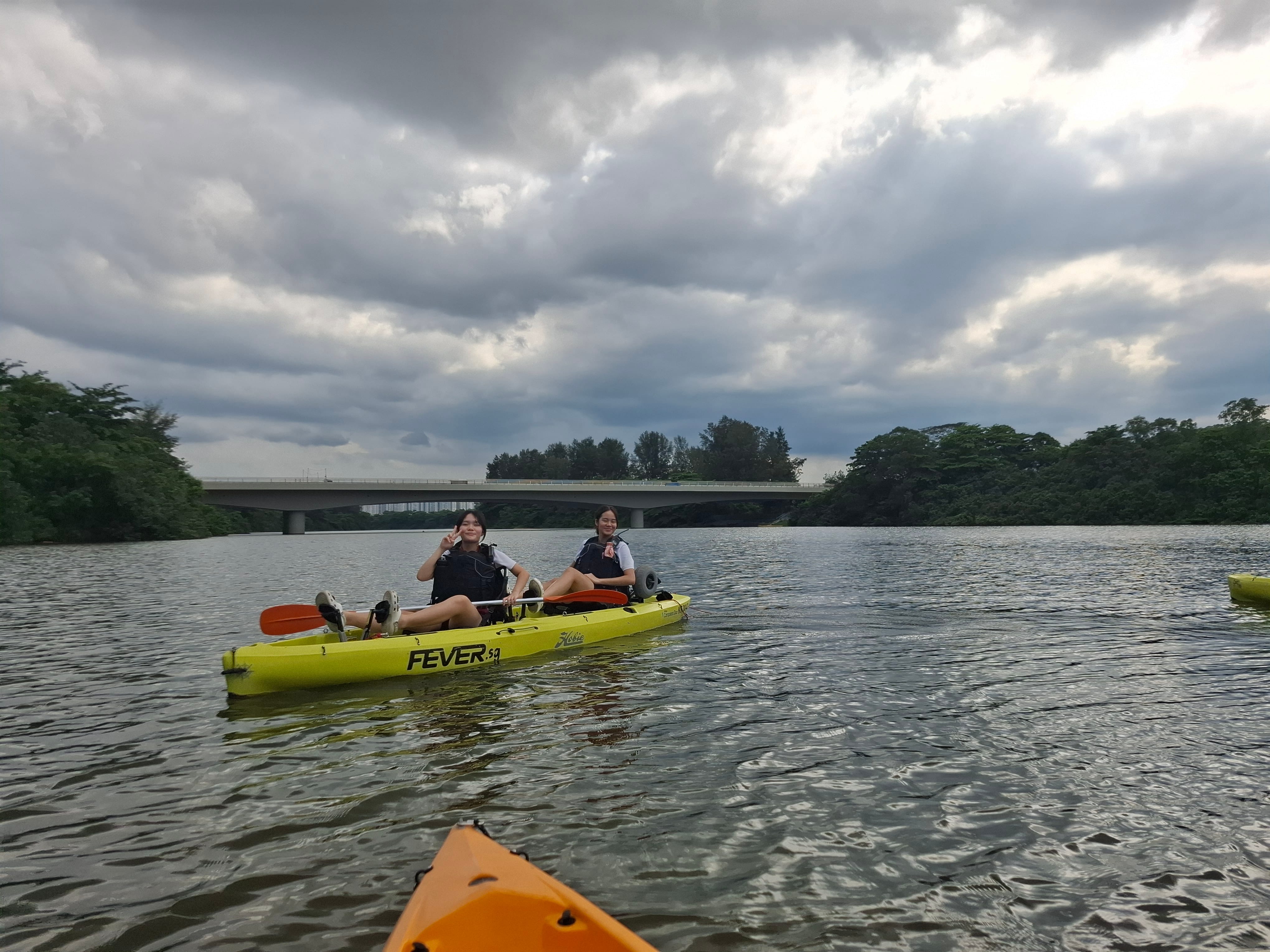 Two people in a yellow kayak on a river, with a bridge behind and dark clouds overhead.