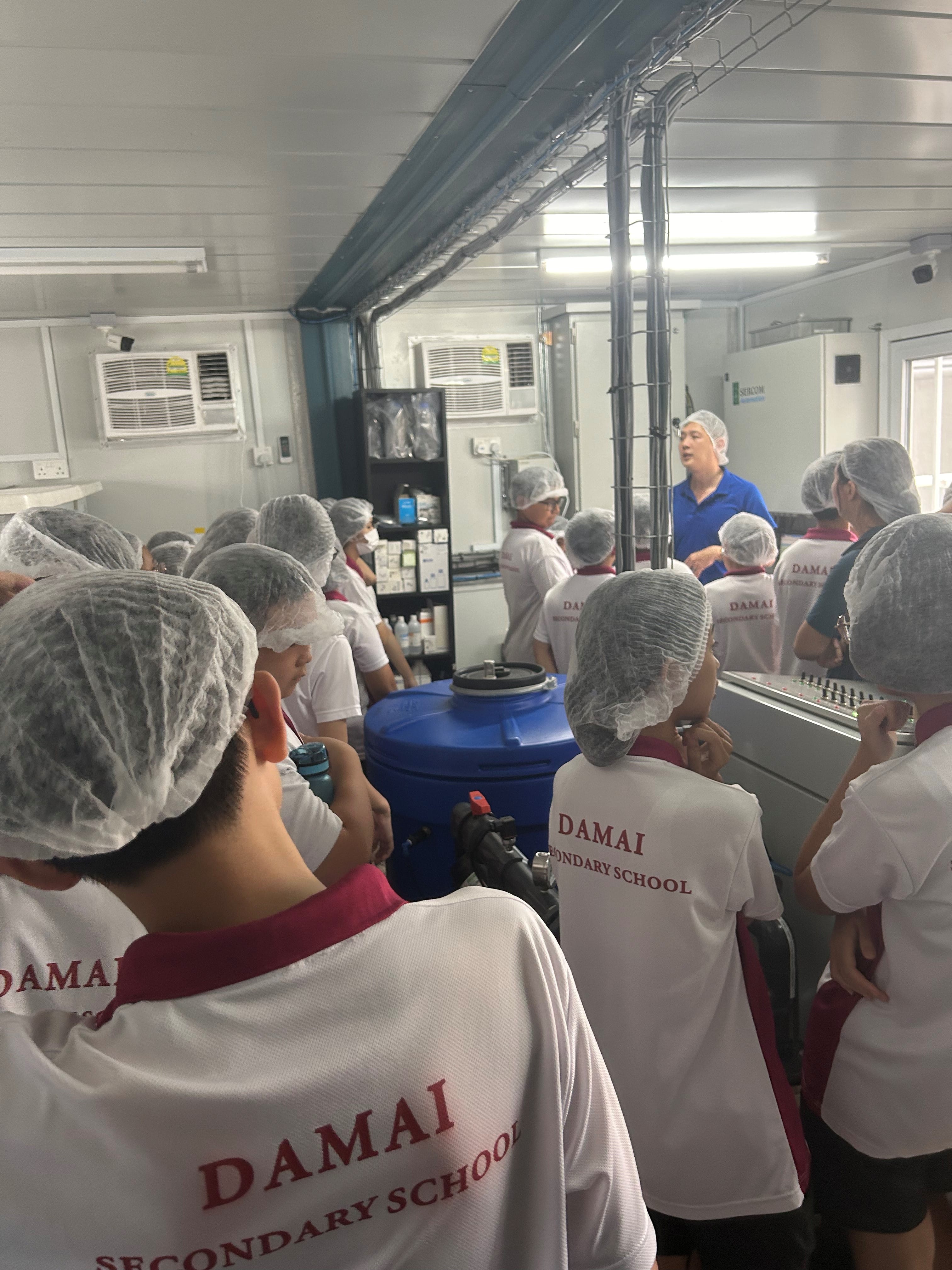 Students in hairnets crowd around a man speaking in a lab-like room with equipment and a blue tank.