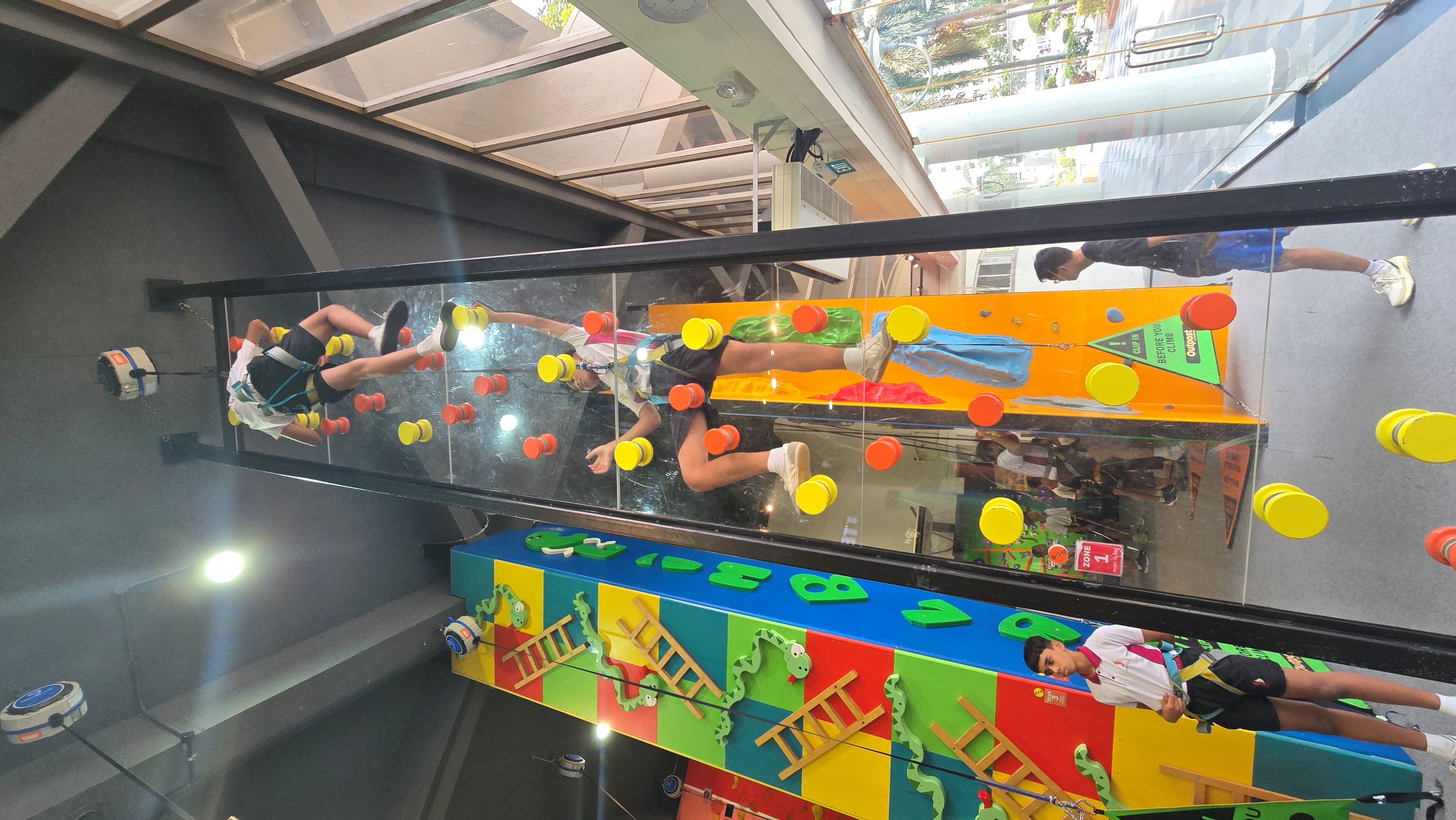 Two children in harnesses climb a glass wall with yellow and orange grips in an indoor activity area.