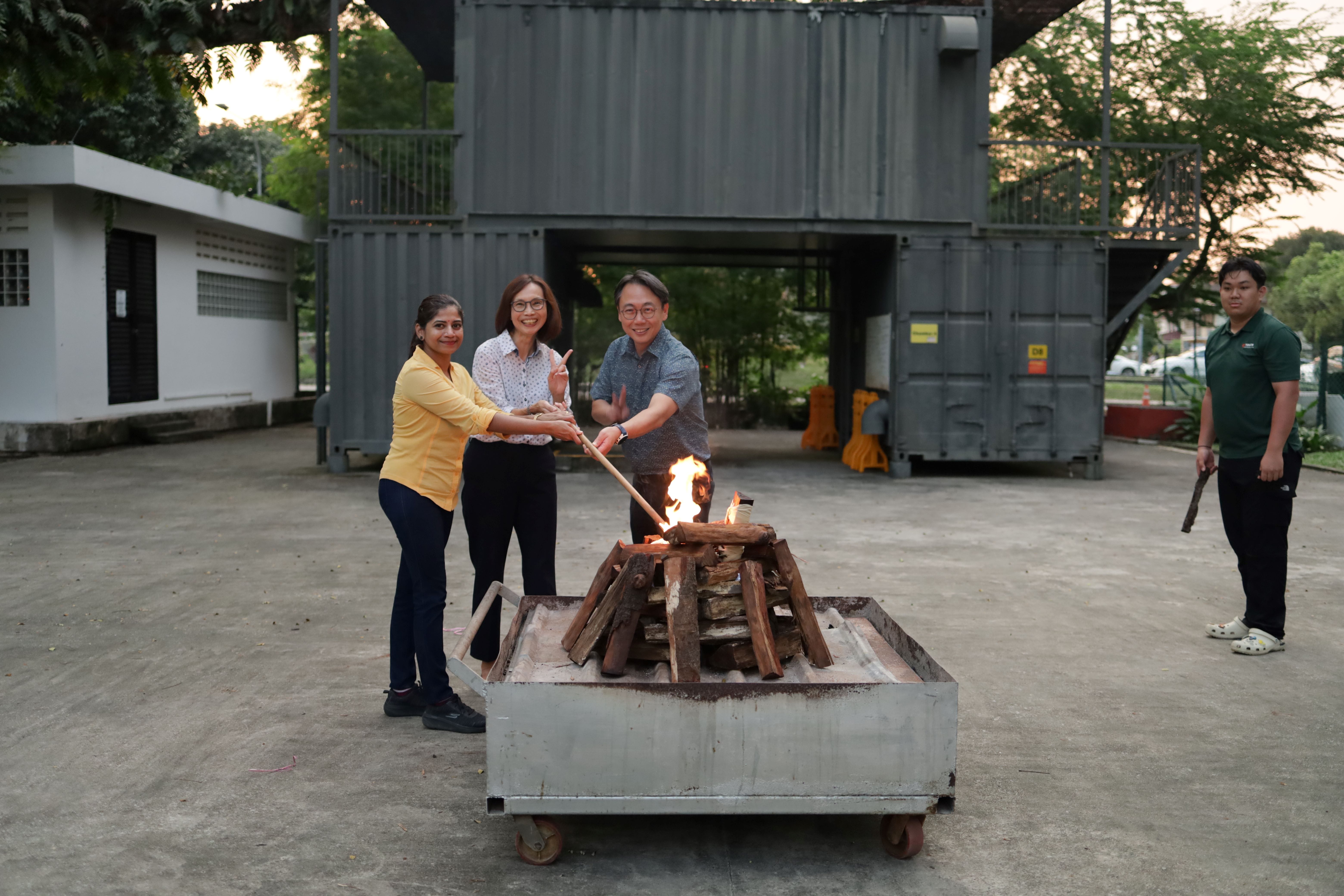 Three people smile while lighting a wood bonfire in a metal cart; another person stands nearby outdoors.