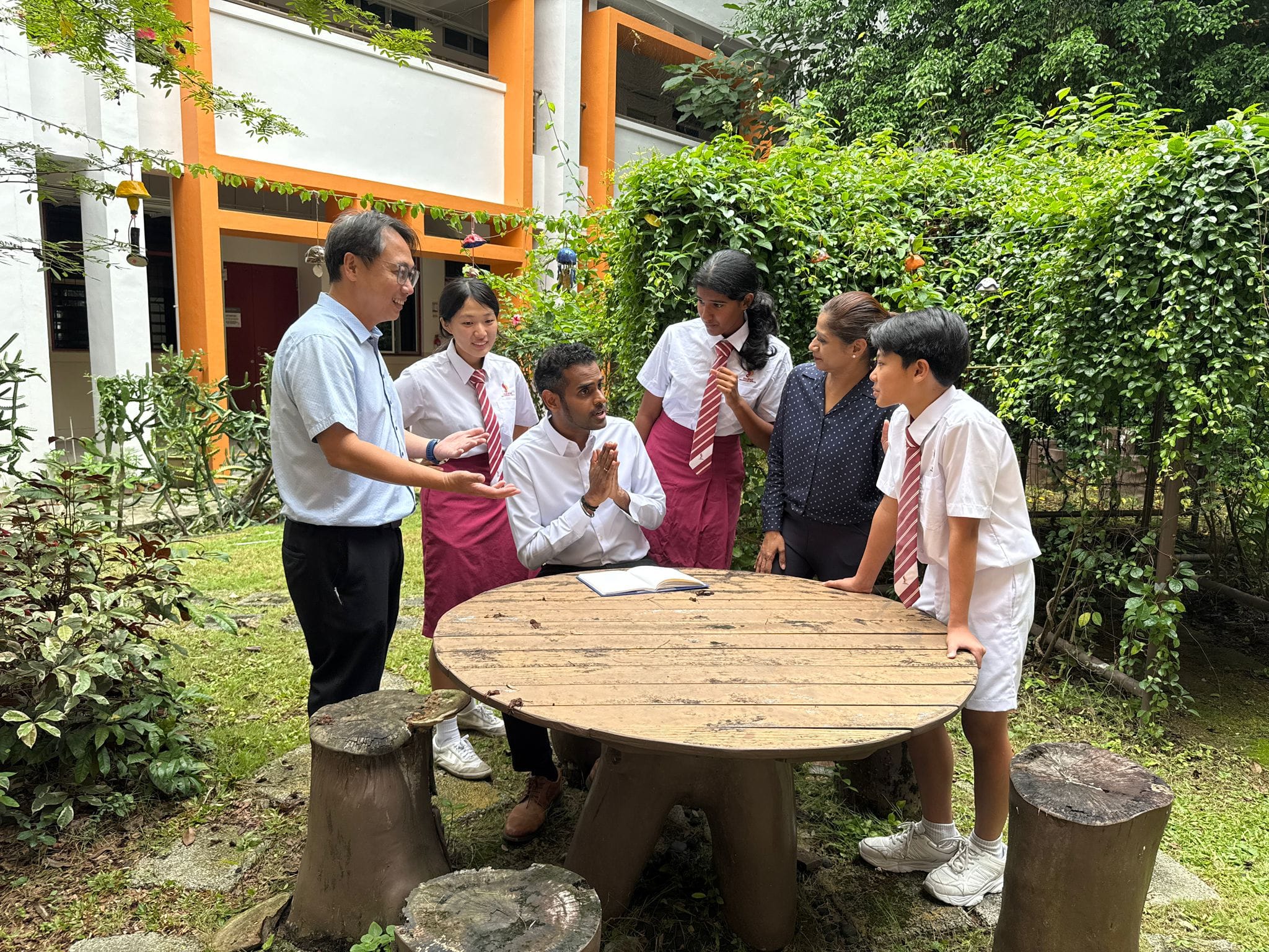 Adults and uniformed students gather around a wooden outdoor table in a school garden, engaged in discussion.