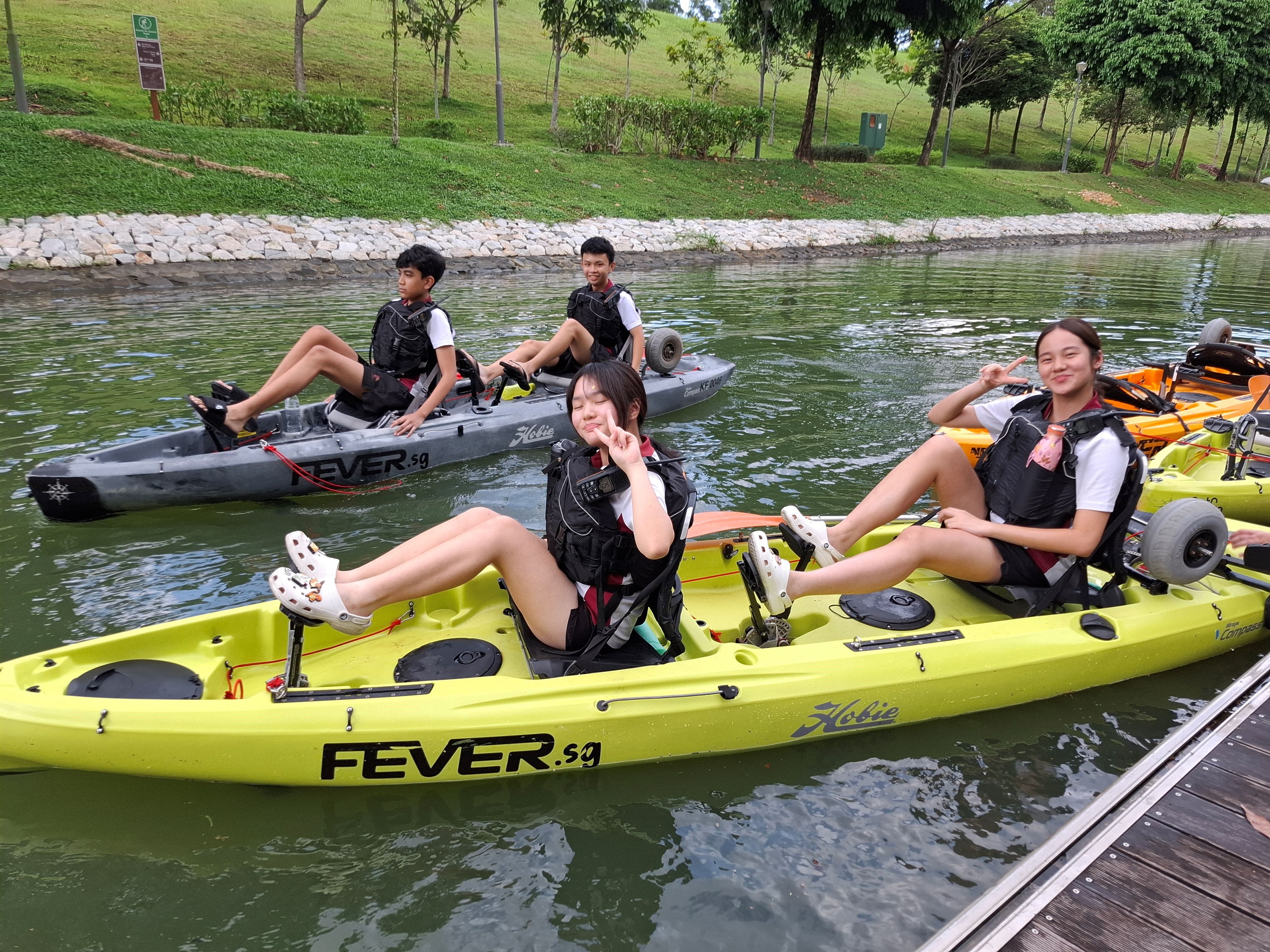 Four teens in life vests relax in kayaks on a calm canal beside a grassy bank, two flashing peace signs.