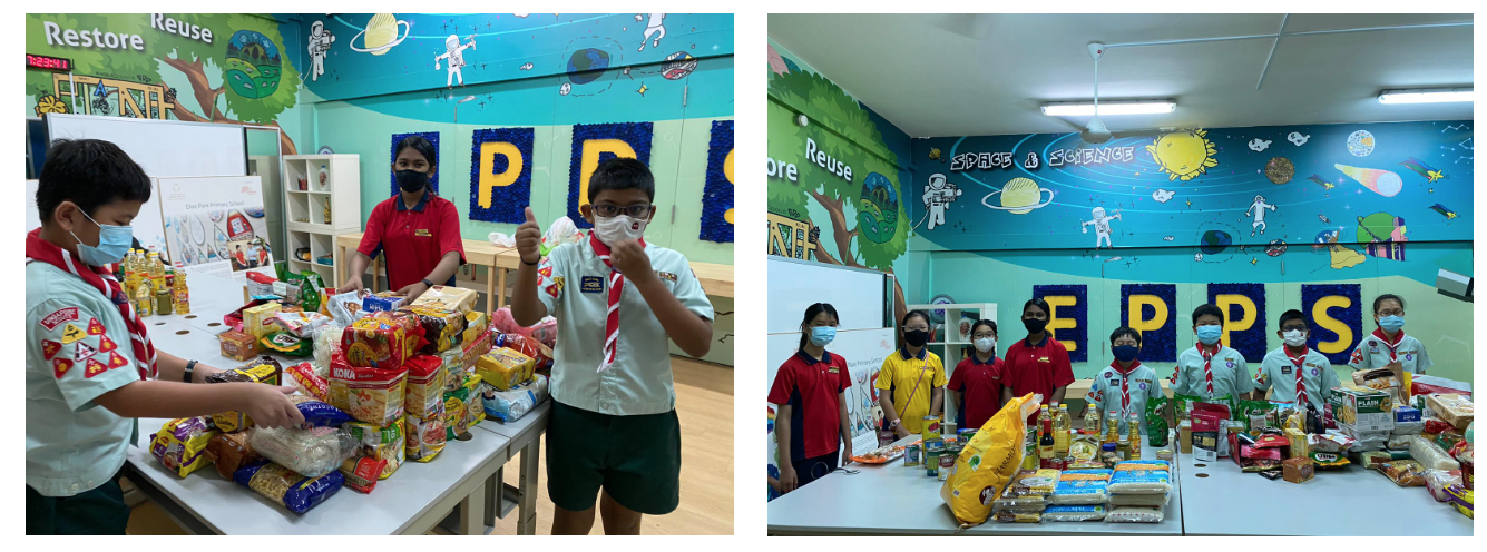 Students in masks and scout uniforms stand beside tables piled with donated food items in a colorful classroom.