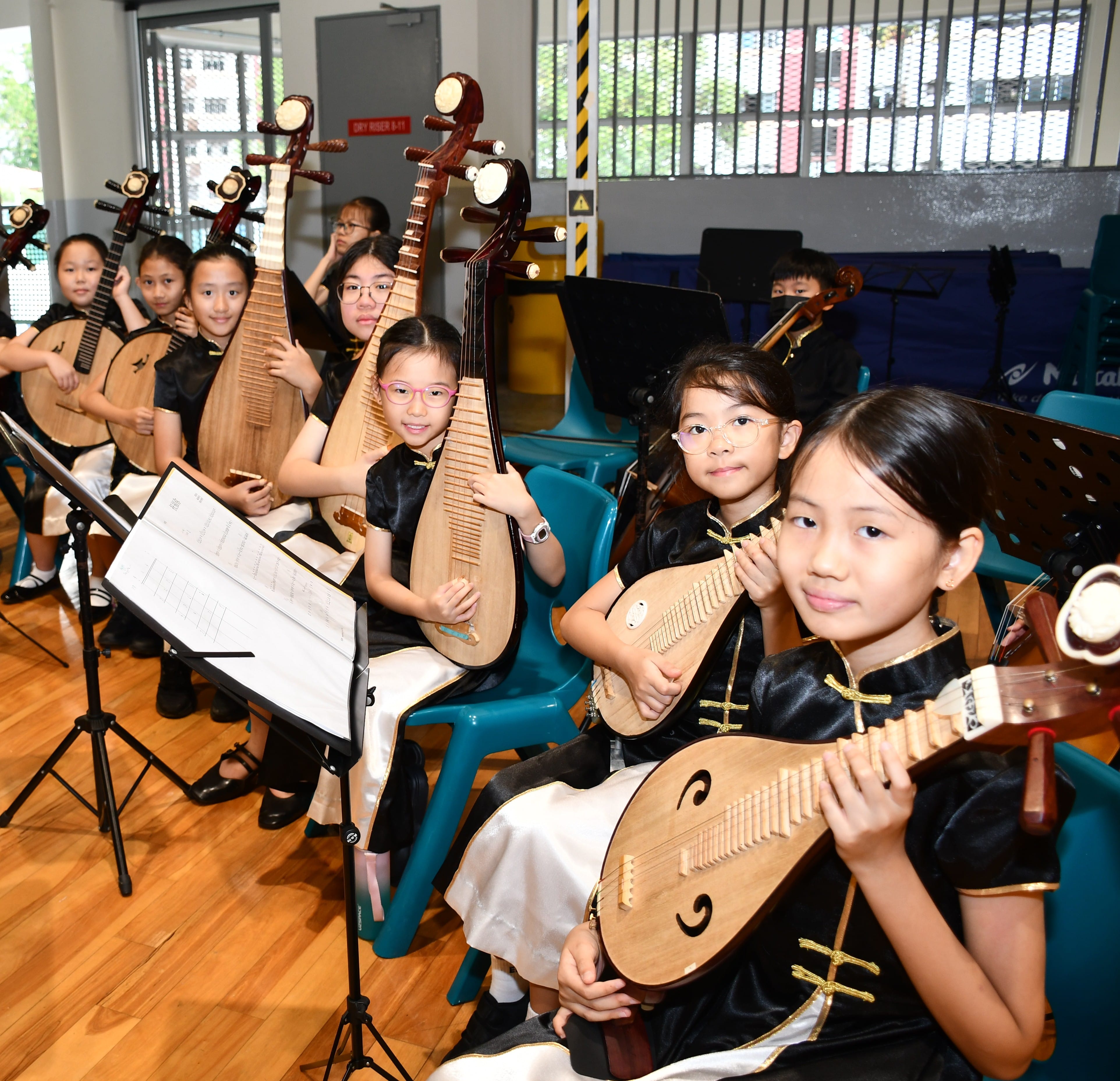A group of young students in matching black-and-white performance outfits smile while seated in a row, holding traditional Chinese stringed instruments like the ruan and pipa during a rehearsal.