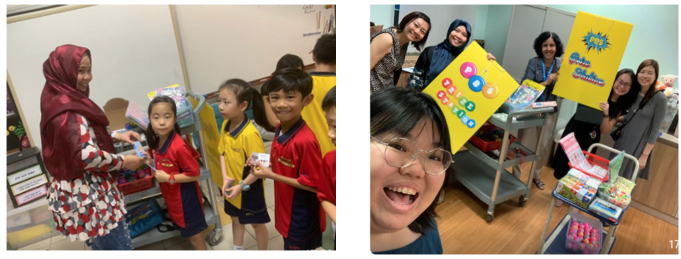 A teacher distributes items to students in uniforms; adults pose with a yellow PBS Prize Station sign and prizes.