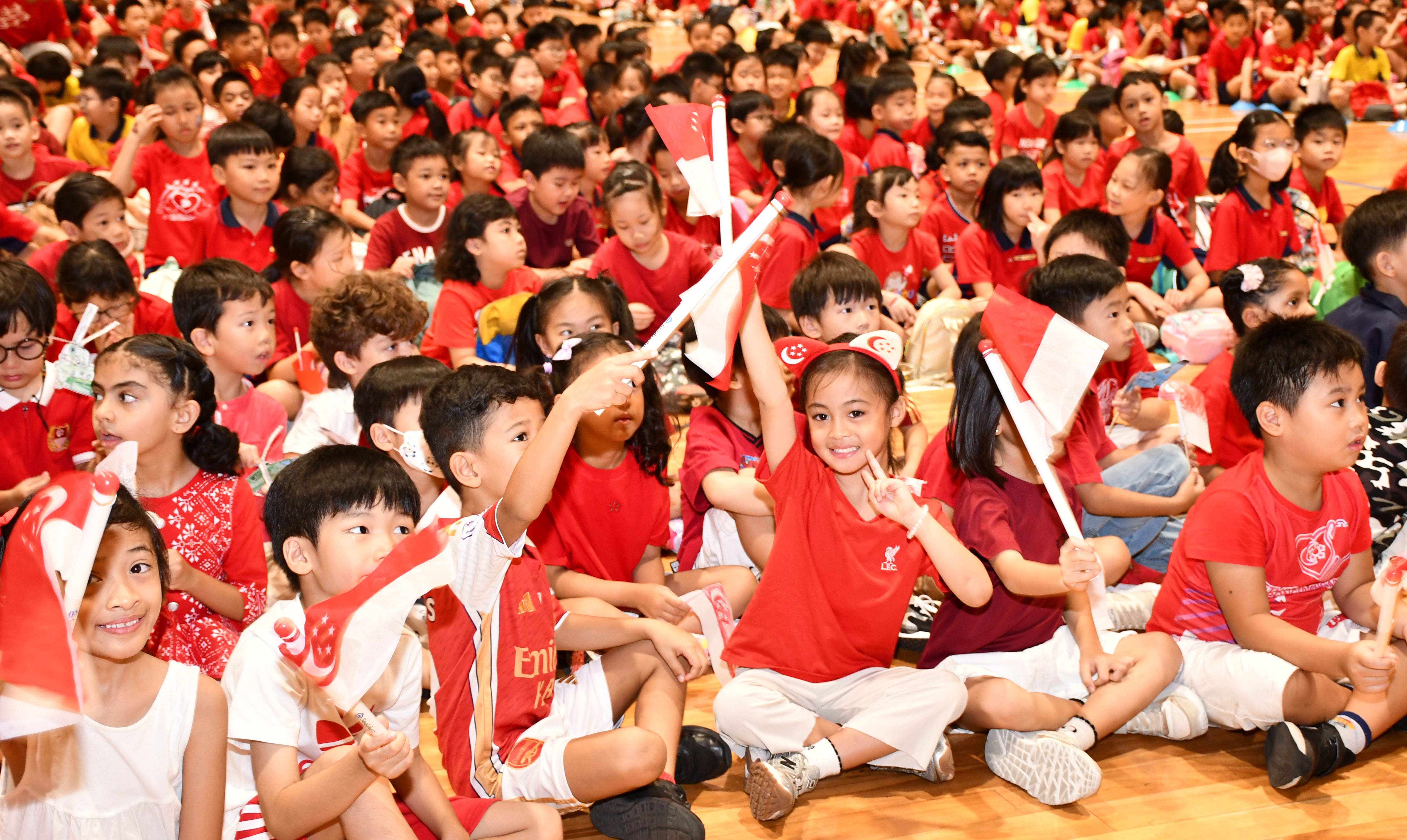 Primary school students dressed in red and white waving handheld Singapore flags during a school National Day celebration in the hall.