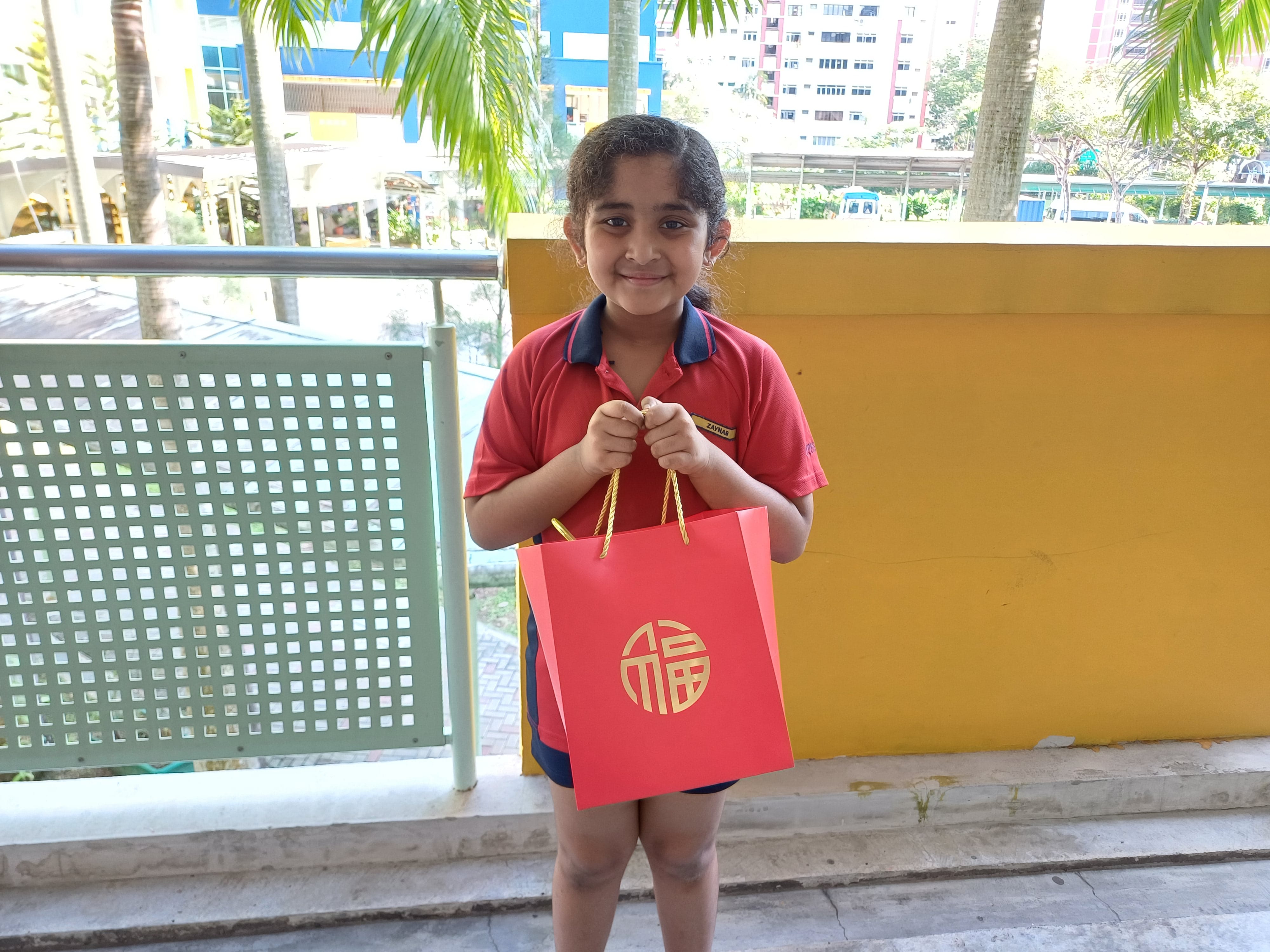 A smiling young student proudly holds a festive red gift bag featuring a golden Chinese character for good fortune.