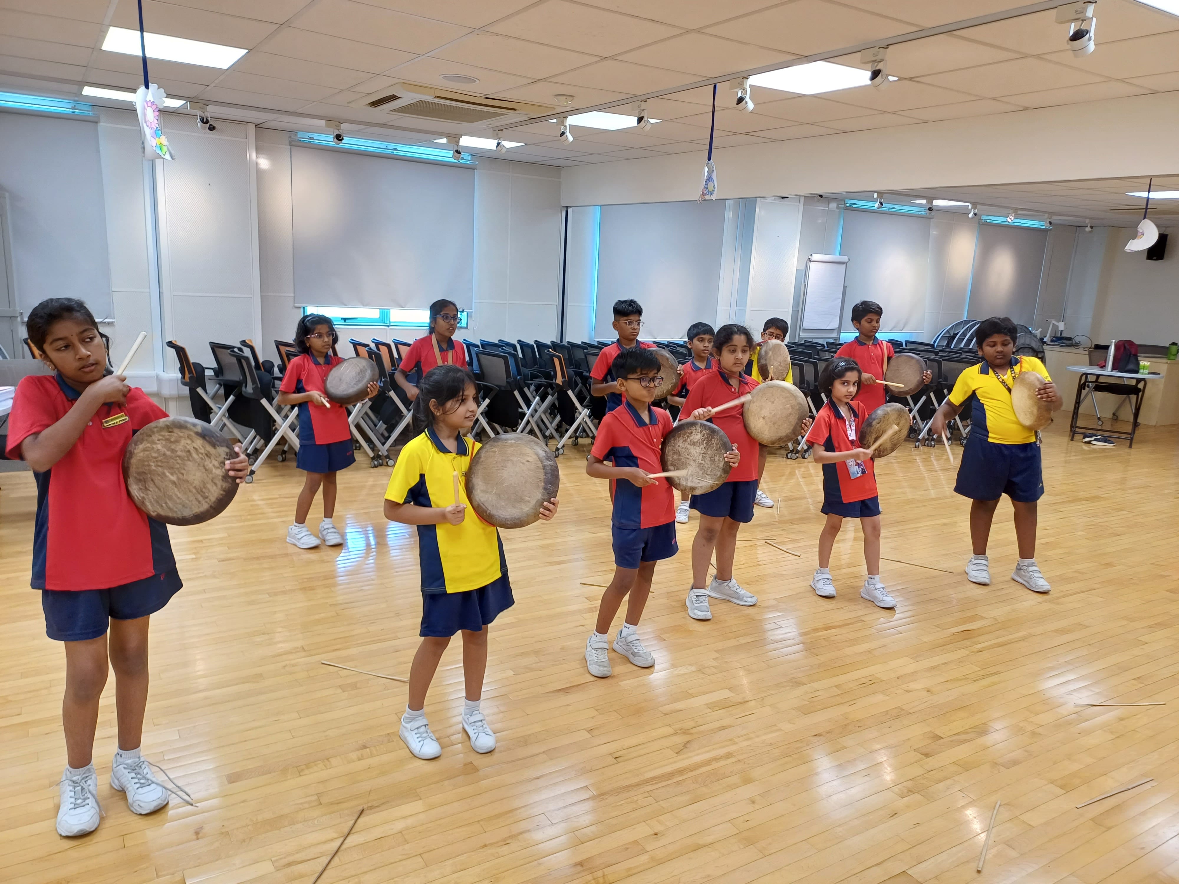 A group of young school children wearing red and yellow polo uniforms standing in an indoor studio space, practicing playing circular frame drums with wooden sticks.