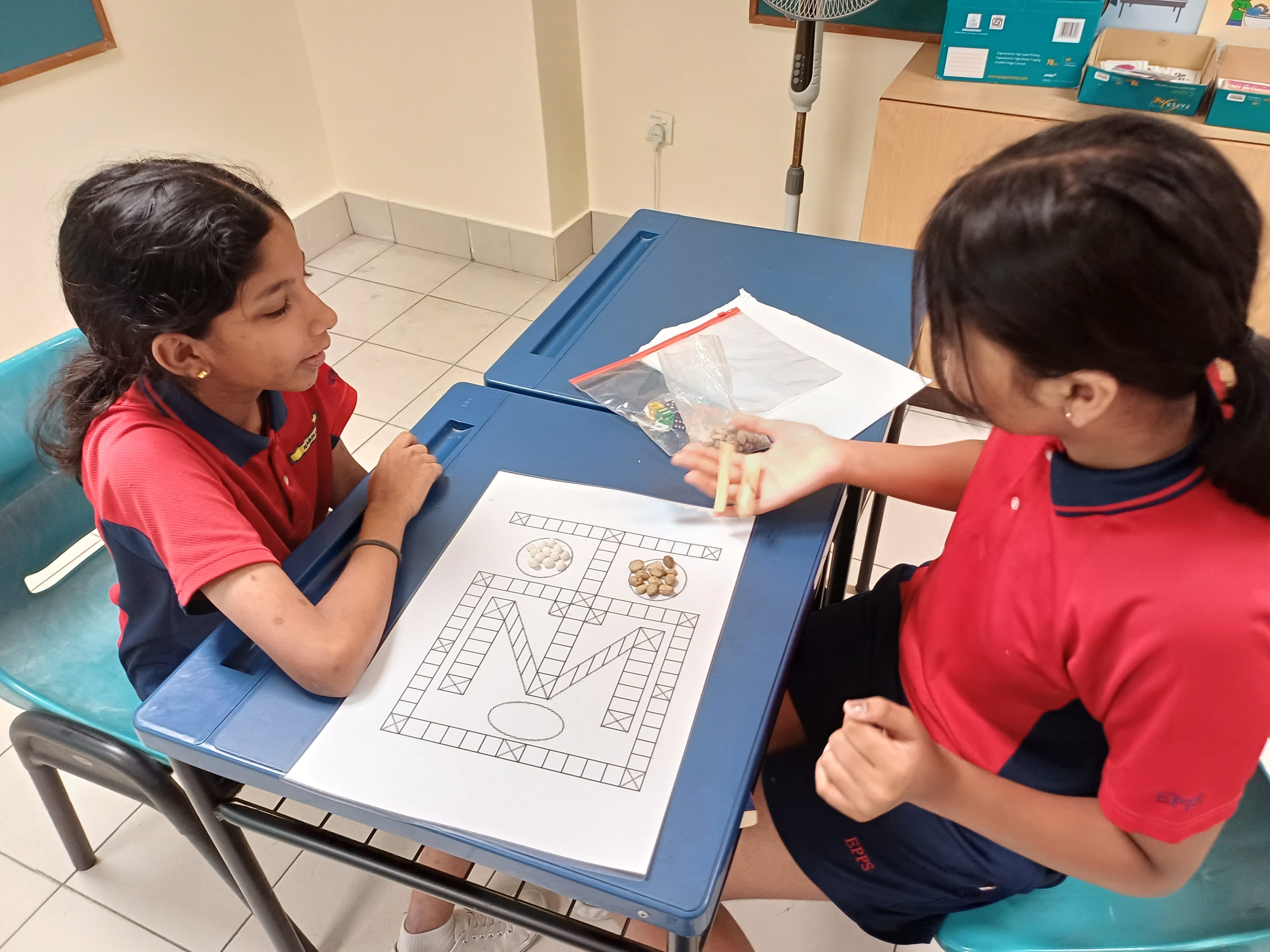 Two young girls in red and navy school uniforms sitting across a blue desk, playing a printed traditional Indian board game with cowrie shells and small sticks.