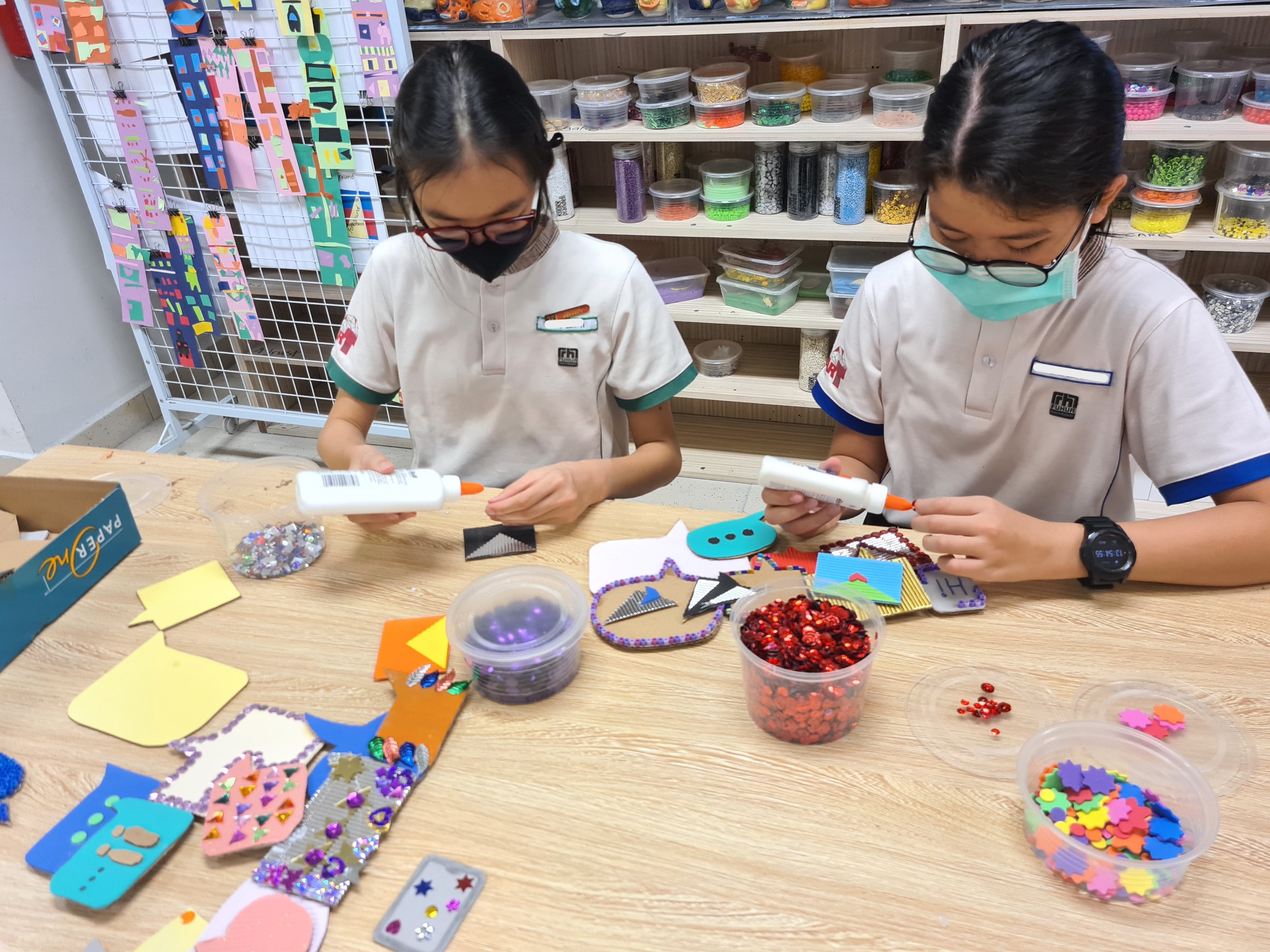 Two masked students in uniforms use glue guns to decorate crafts with sequins and colorful paper at a table.