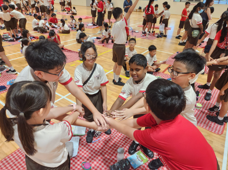 A group of school children in uniforms stacking hands together in a gym filled with seated students.
