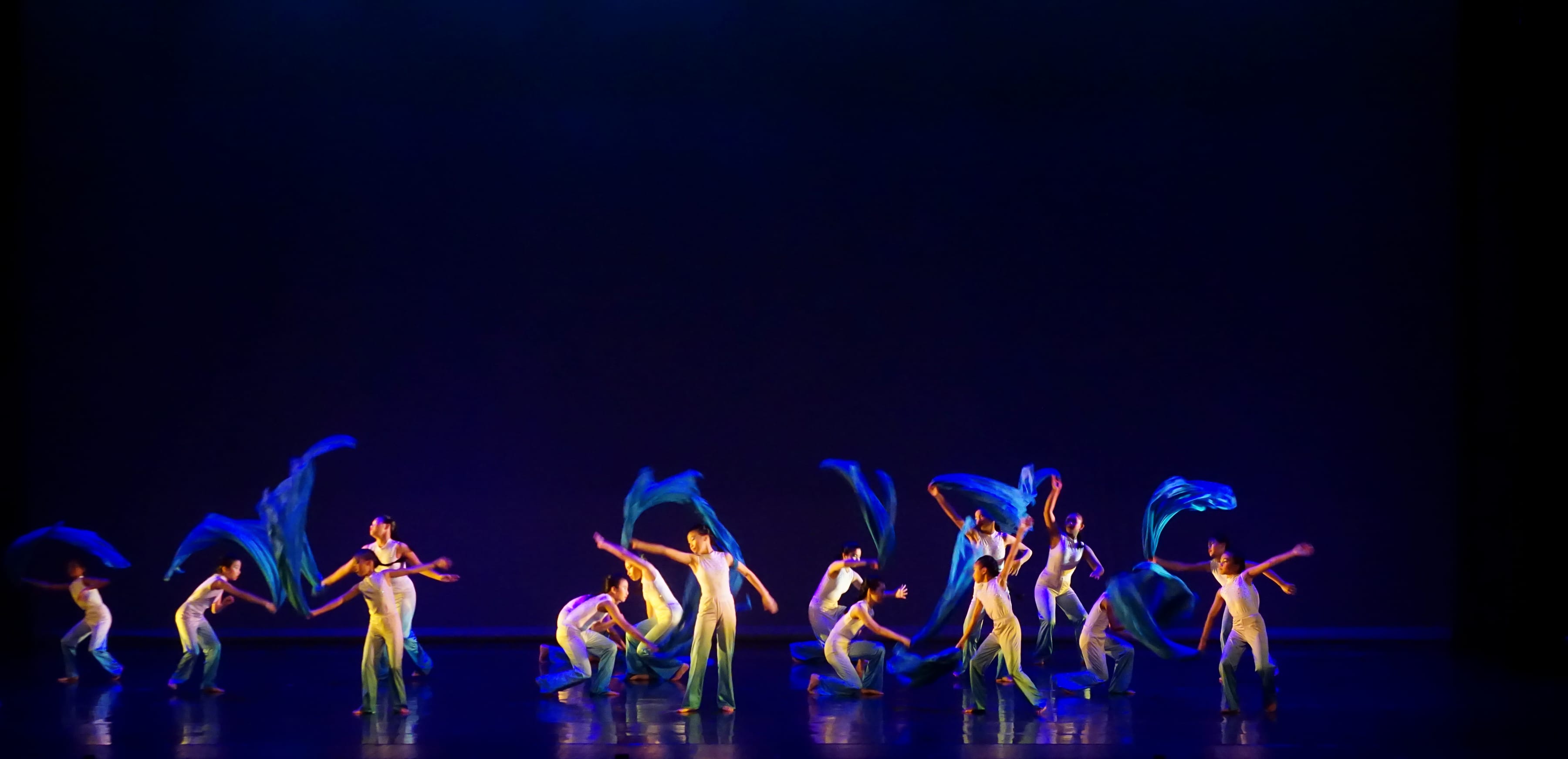 Dancers in white costumes wave flowing blue fabric on a dark stage lit with blue and warm light.