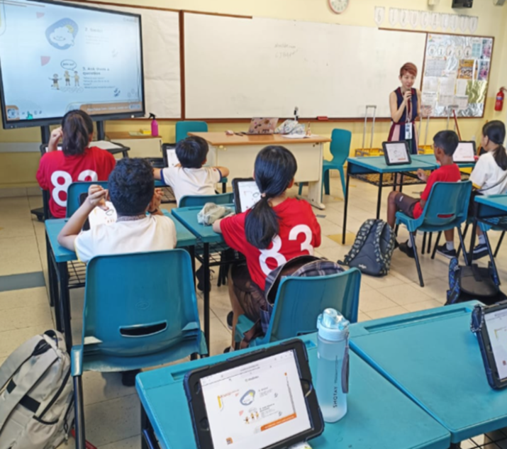Teacher stands at front of classroom while students sit at teal desks with tablets, facing a presentation screen.