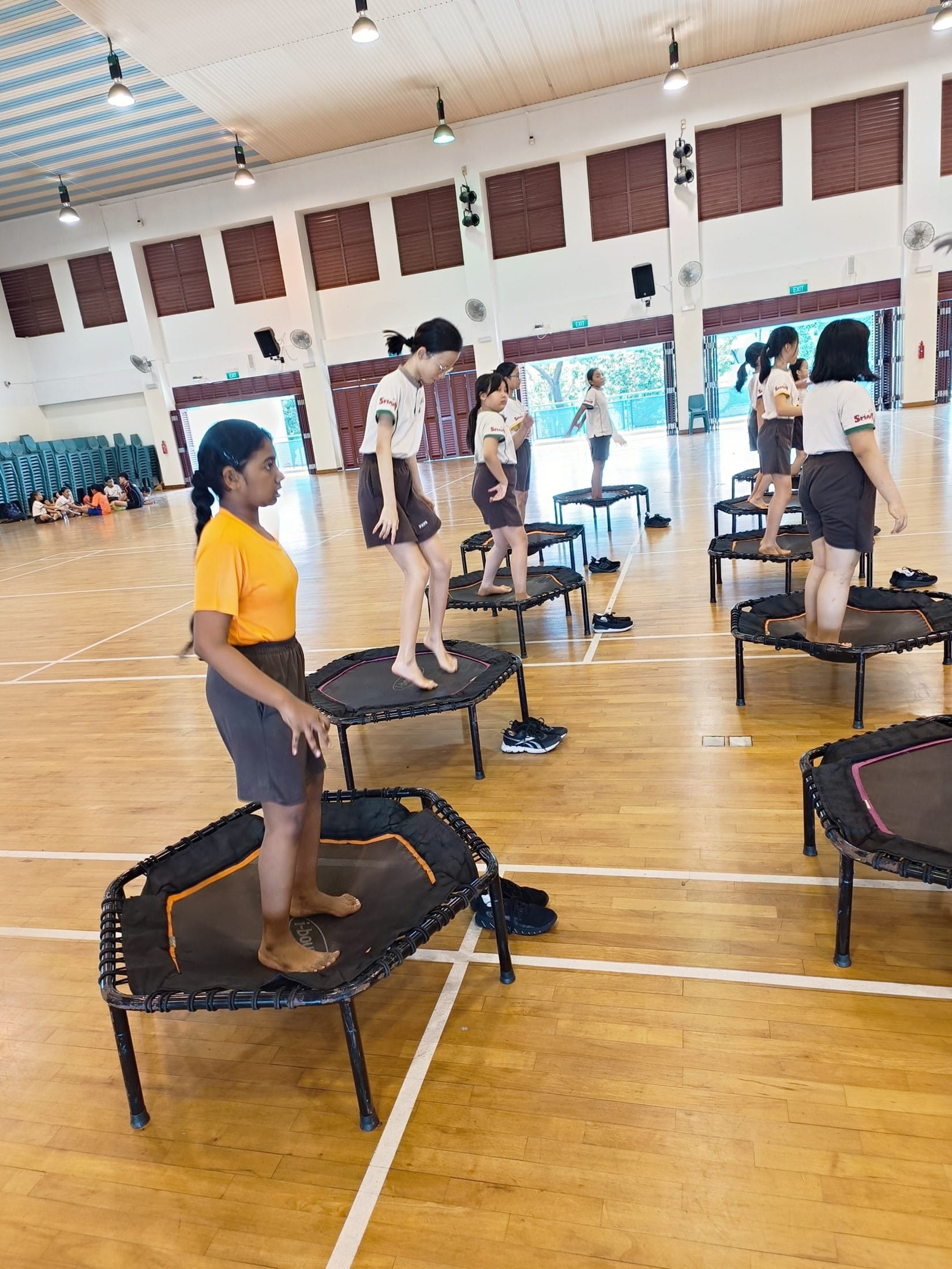 Students in school uniforms stand on small individual trampolines in a gymnasium during a fitness class.