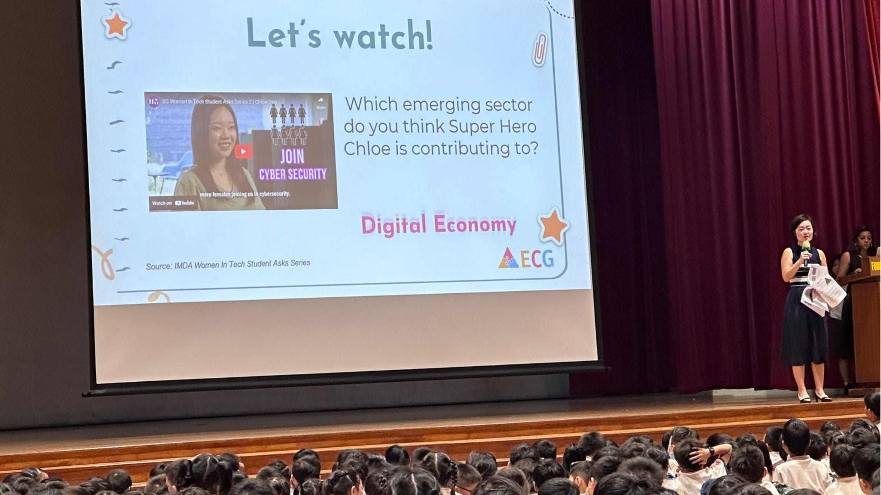 A woman with a microphone presents a slide about Digital Economy and cyber security to a large student audience in a hall.