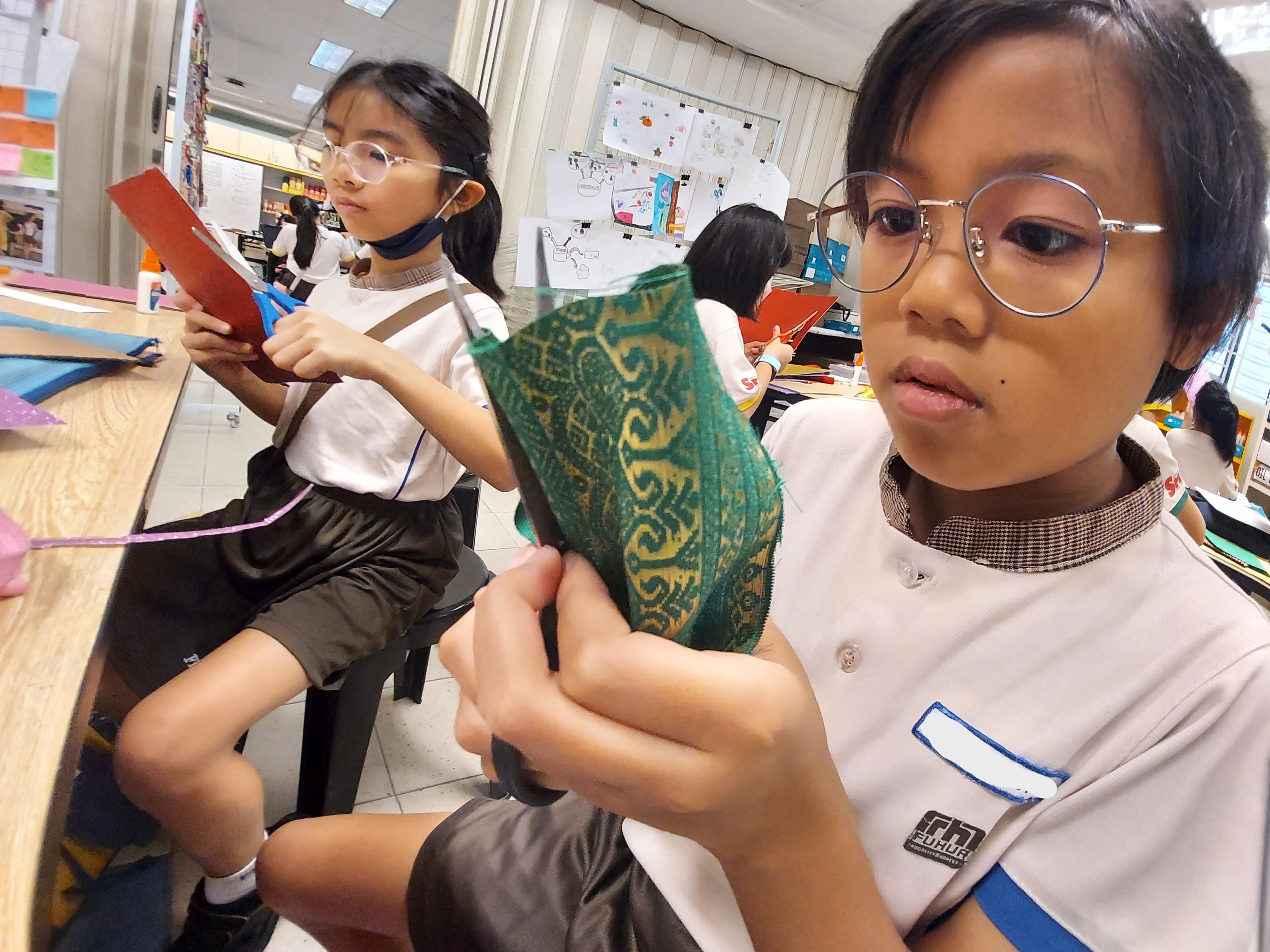 Two young students in school uniforms cut patterned fabric and red paper with scissors in a classroom.