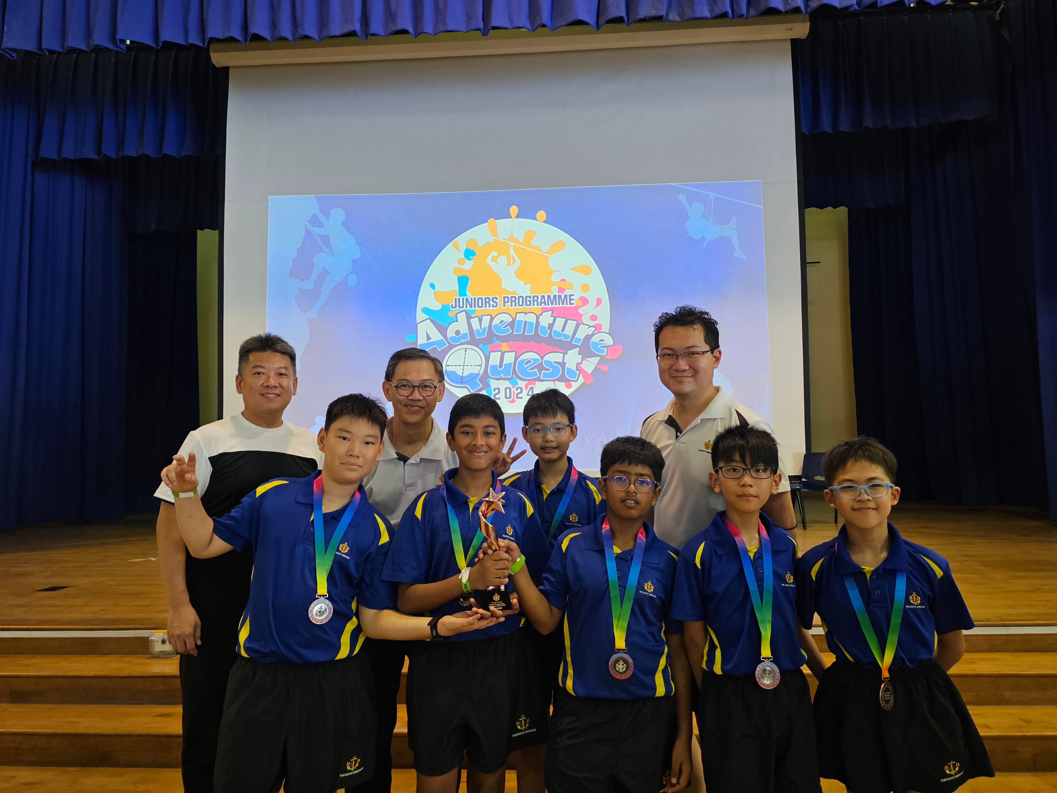 Six students in blue uniforms with medals and a trophy pose with three adults before an Adventure Quest 2024 screen.