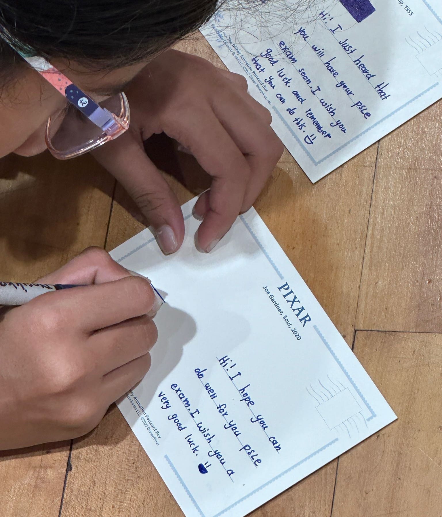 Child with glasses writing a good luck message on a Pixar postcard, with another handwritten postcard nearby.